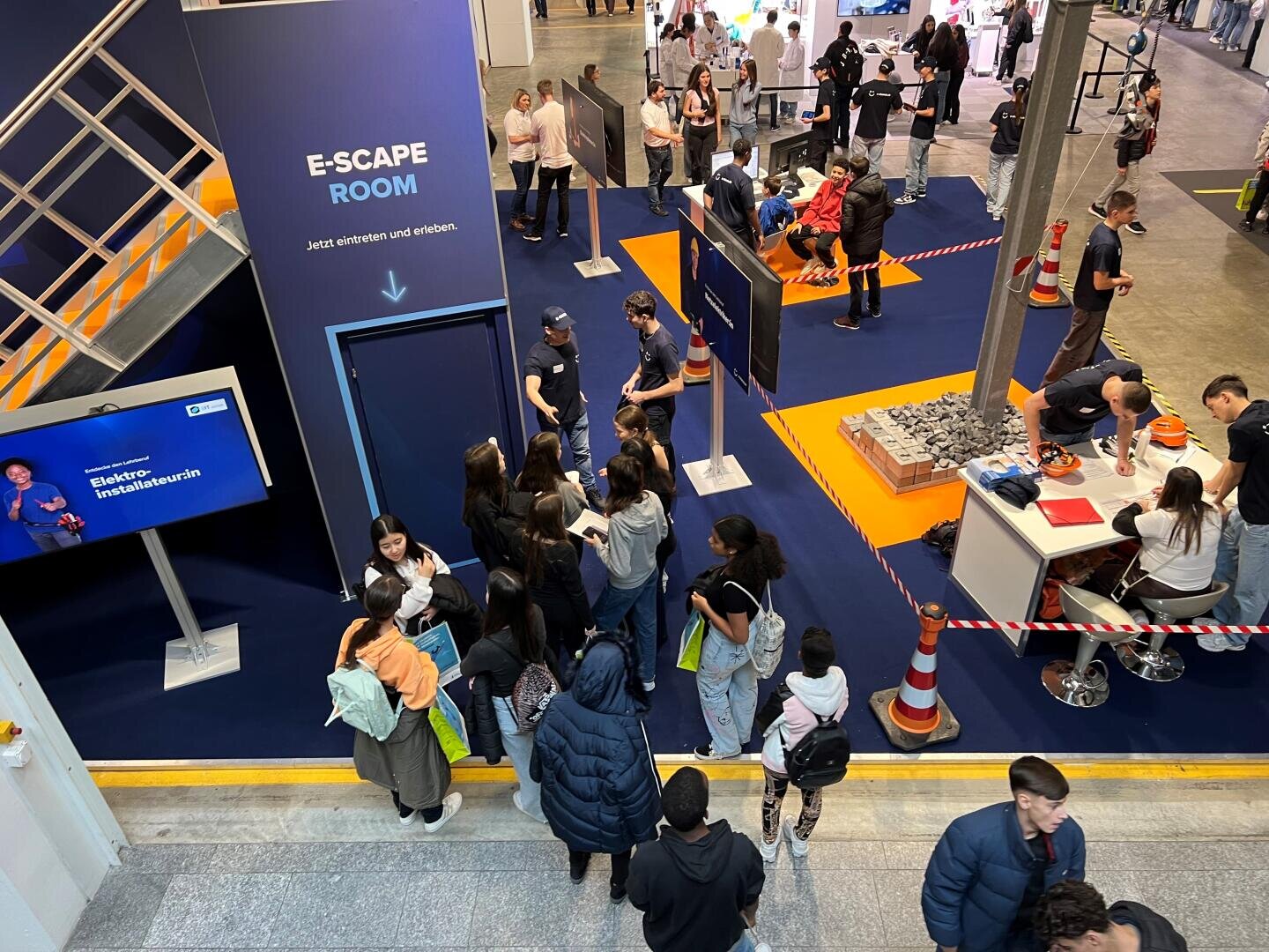 Bei einer Veranstaltung versammeln sich Menschen am Eingang zu einem E-SCAPE-ROOM-Stand. Einige stehen Schlange, andere unterhalten sich in der Nähe. Zu sehen sind ein orange-blauer Bodenbelag, verschiedene Stände und eine Treppe.