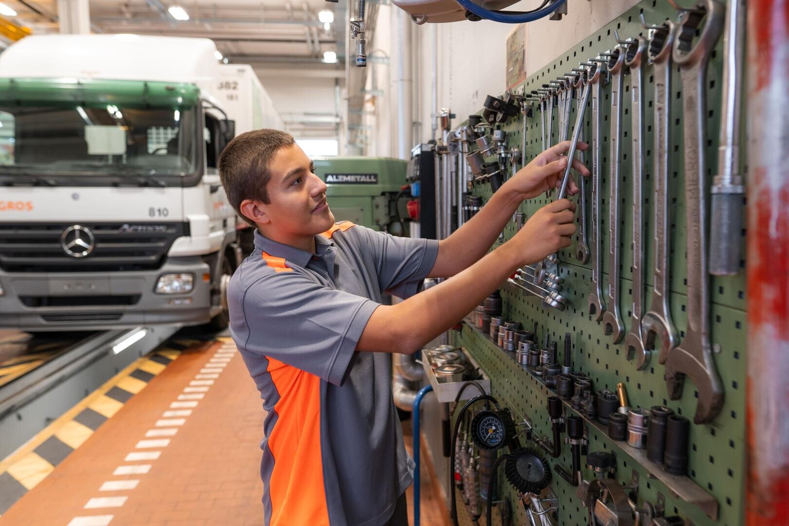 Ein junger Mensch in einem grau-orangefarbenen Hemd nimmt einen Schraubenschlüssel von einer Werkzeugtafel in einer Werkstatt, im Hintergrund ist ein weißer Lieferwagen zu sehen.