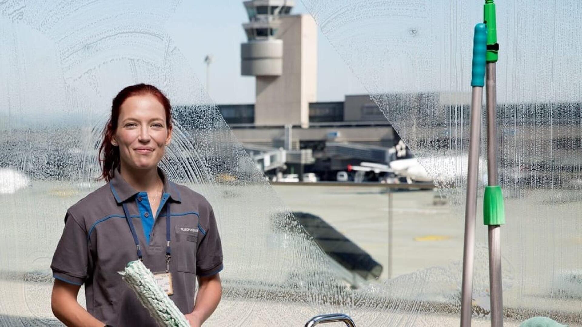 Eine Frau in einer grauen Uniform steht lächelnd da und putzt ein großes Flughafenfenster, während im Hintergrund Flugzeuge und ein Flugsicherungsturm zu sehen sind. Im Vordergrund sind Reinigungswerkzeuge zu sehen.