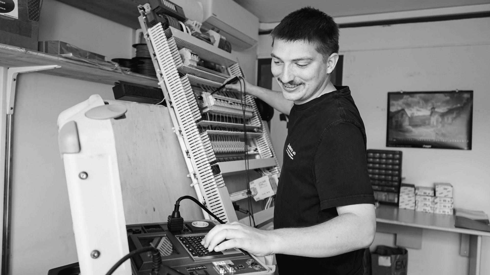 Ein Mann lächelt, während er in einer Werkstatt mit elektronischen Geräten arbeitet, ein Bedienfeld bedient und eine große, offene Leiterplatte an der Wand untersucht. Im Hintergrund befinden sich Regale mit Kisten und ein gerahmtes Bild.