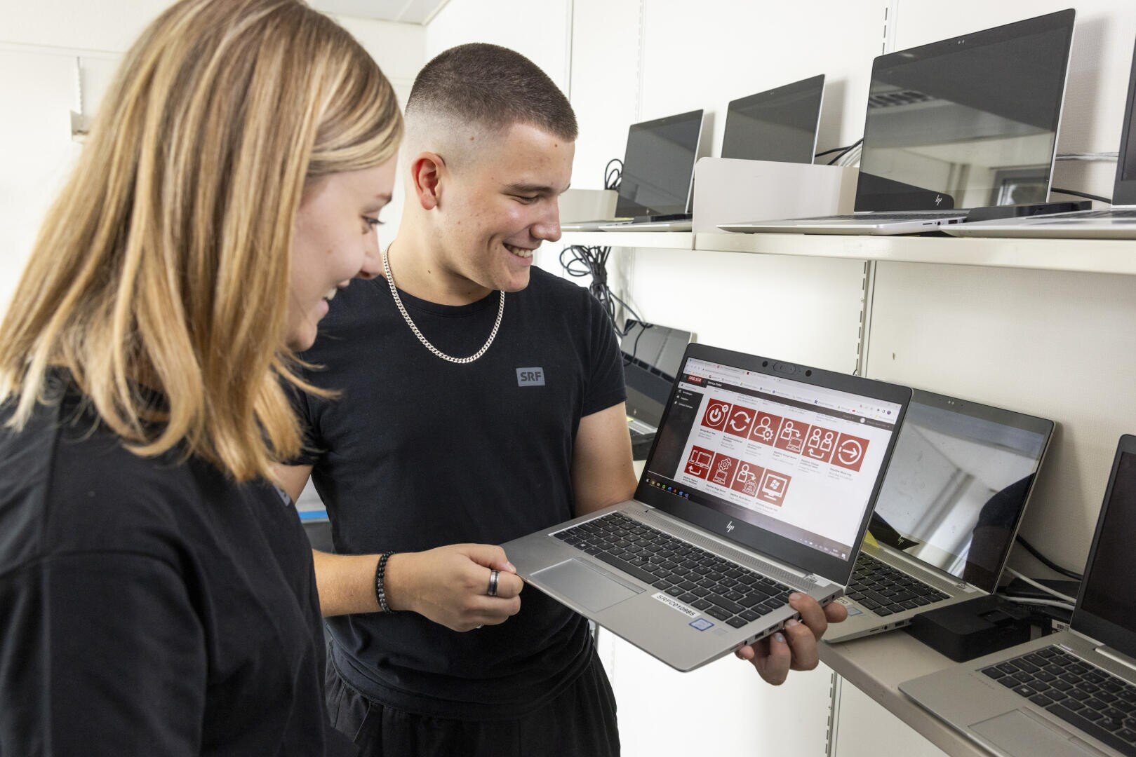 Zwei junge Menschen stehen vor Regalen mit Laptops, lächeln und schauen auf einen Laptop-Bildschirm, auf dem verschiedene rote und weiße Symbole oder Grafiken zu sehen sind.