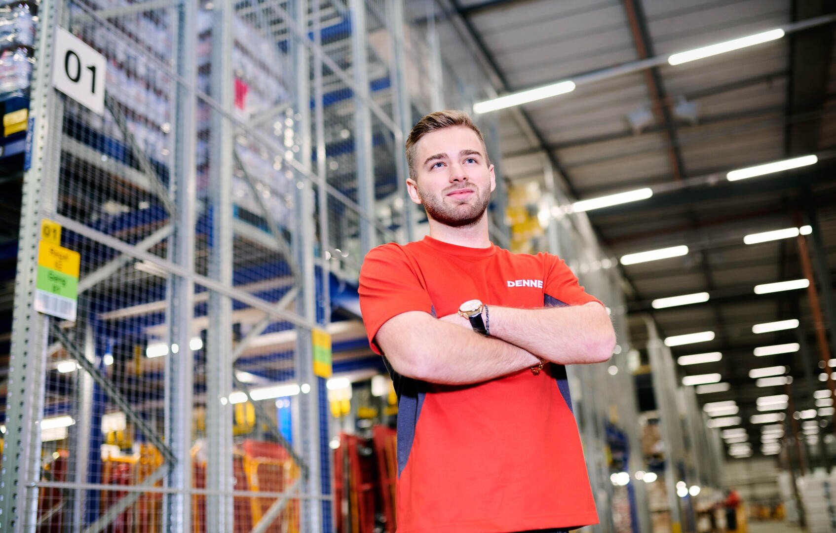 Ein Mann in einem roten Hemd steht mit verschränkten Armen in einer großen Lagerhalle, umgeben von hohen Regalen, die mit Kartons und Waren gefüllt sind. Die Beleuchtung ist hell und im industriellen Stil gehalten.