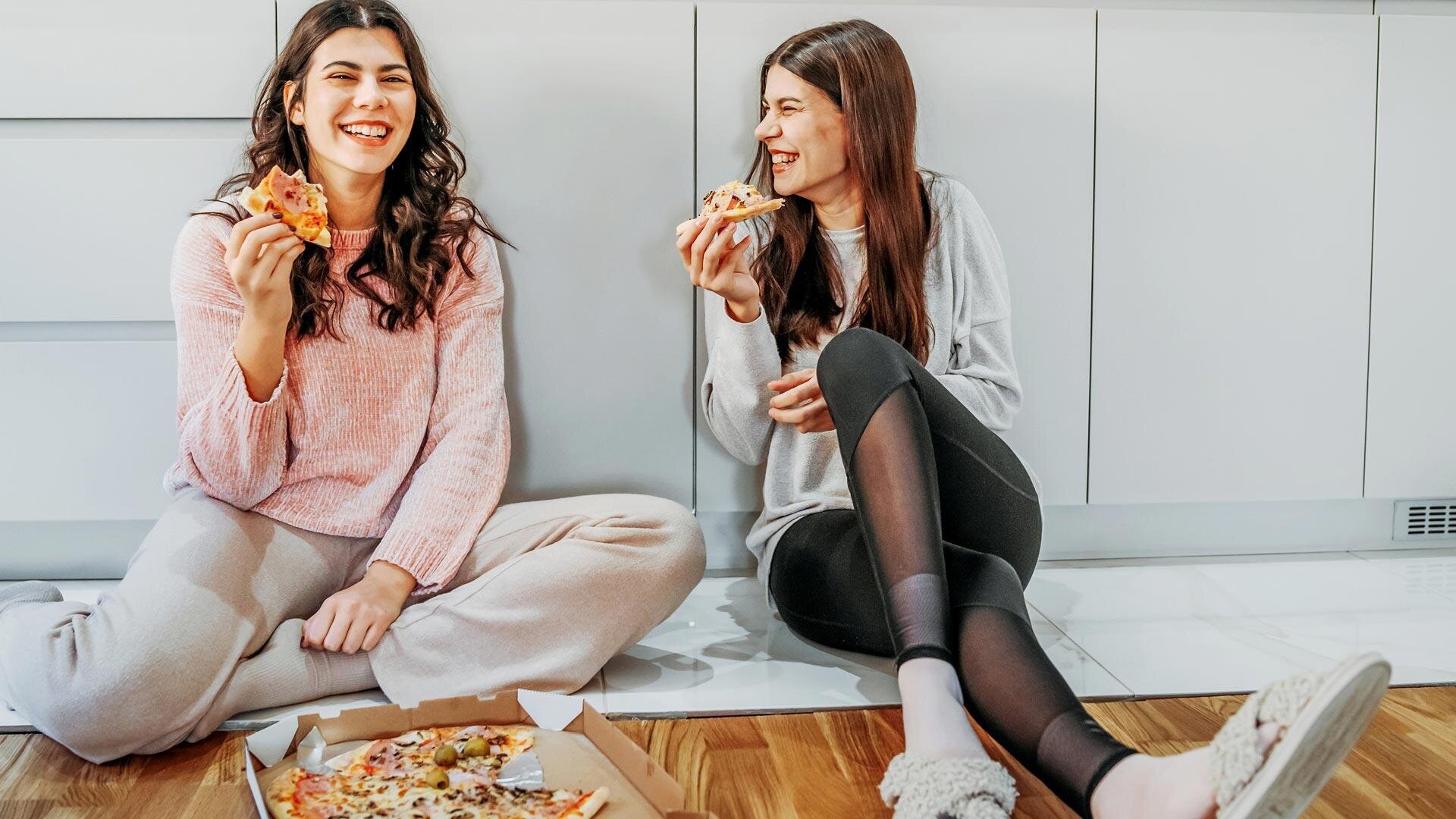 Zwei Frauen sitzen auf dem Boden in einer Küche, lächeln und essen Pizza aus einer offenen Schachtel. Sie wirken entspannt und glücklich und genießen ihr Essen und die gemeinsame Zeit.