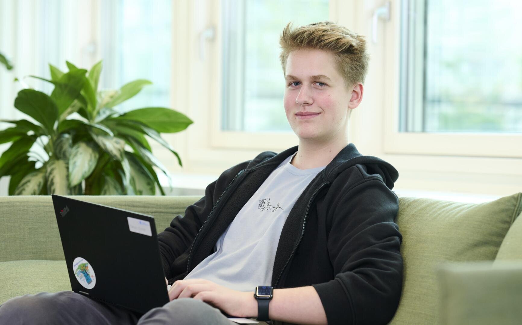 Ein junger Mensch mit kurzen blonden Haaren sitzt auf einem grünen Sofa, arbeitet an einem Laptop und lächelt leicht. Im Hintergrund sind große Fenster und eine Blattpflanze zu sehen.
