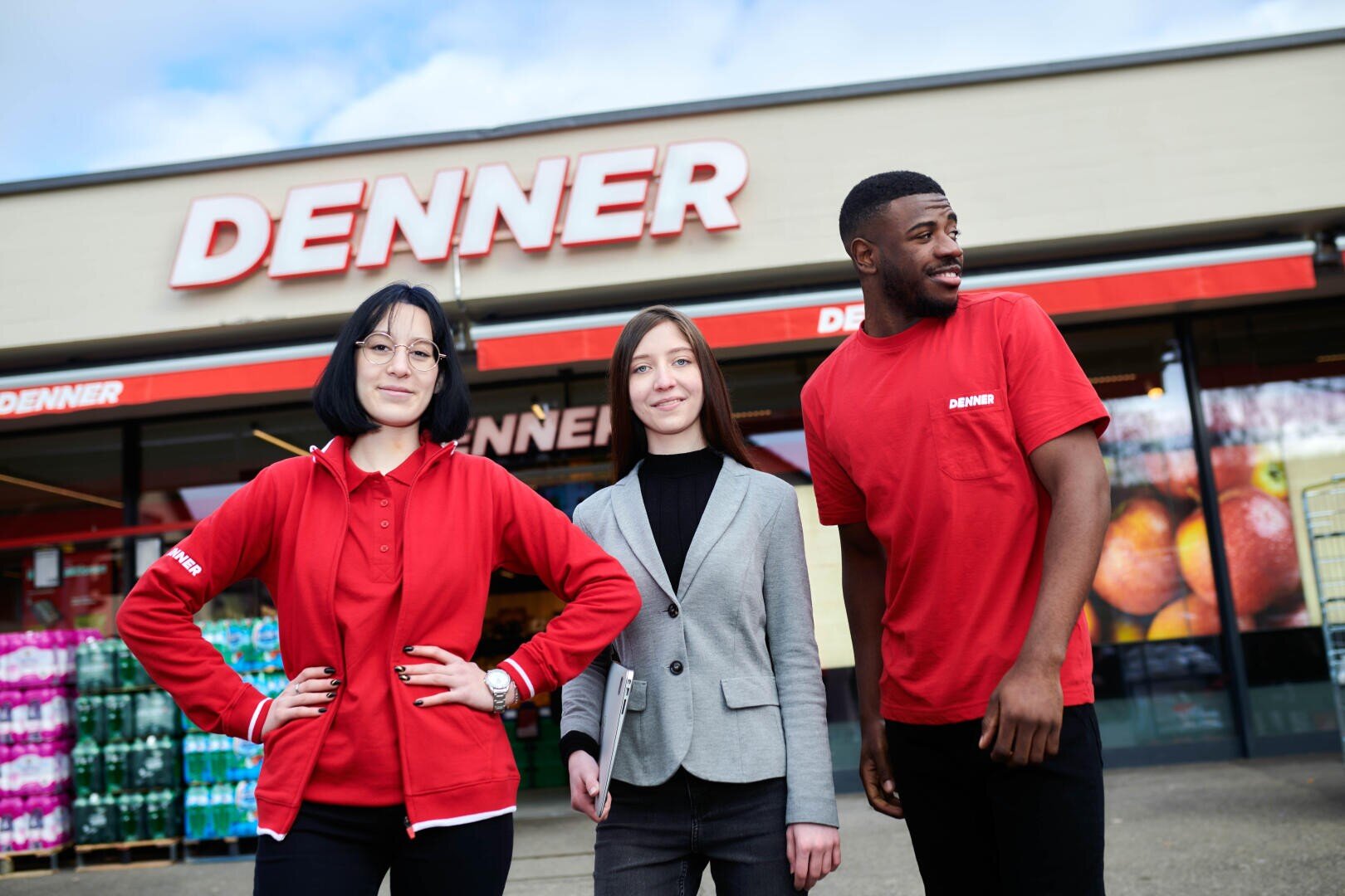 Drei Personen stehen lächelnd vor einem Denner-Geschäft. Zwei tragen rote Denner-Uniformen, eine trägt einen grauen Blazer. Der Name des Geschäfts ist auf dem Gebäude hinter ihnen unter einem teilweise bewölkten Himmel zu sehen.