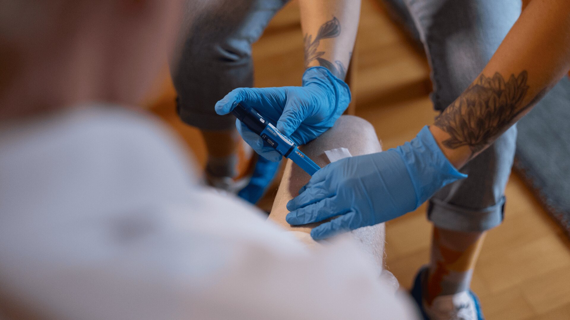 Eine Person mit blauen Handschuhen benutzt ein Blutzuckermessgerät am Arm einer anderen Person und prüft deren Blutzuckerwerte. Der Tester hat Tätowierungen auf den Armen, und die Umgebung scheint ein Innenraum mit Holzboden zu sein.