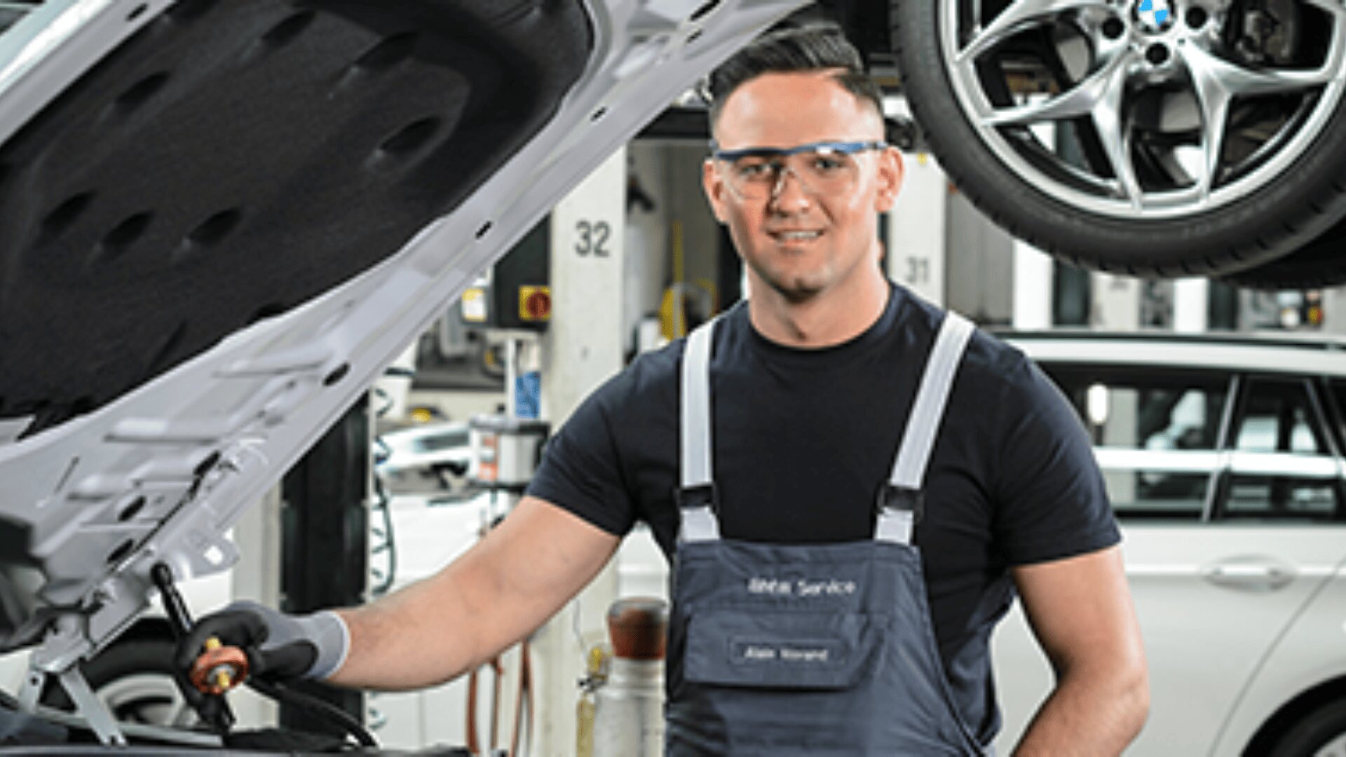 Ein Mechaniker mit Schutzbrille und dunklem Overall steht in einer Garage neben einer offenen Motorhaube und lächelt in die Kamera. Im Hintergrund ist ein Autoreifen zu sehen.