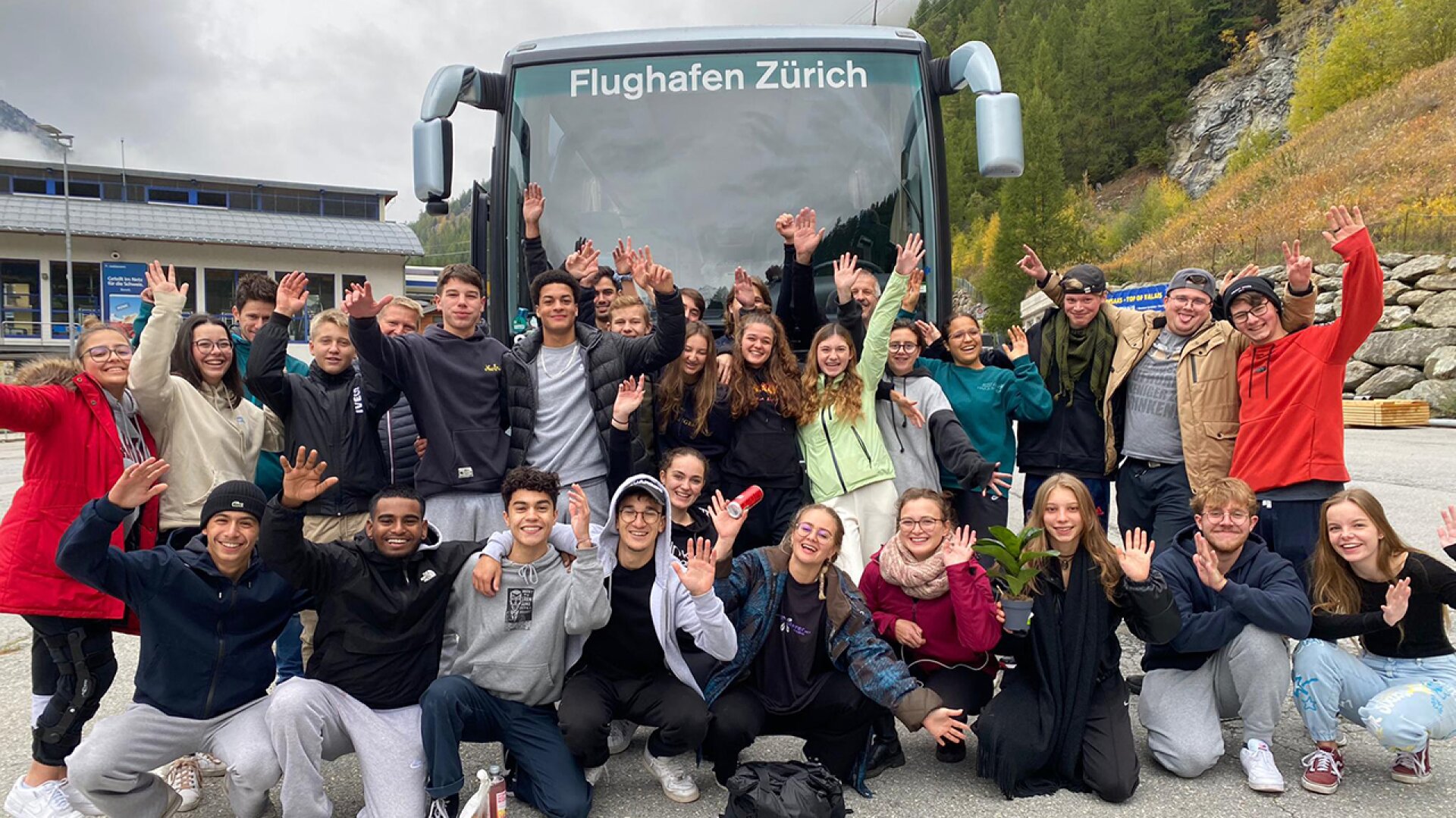 Eine Gruppe lächelnder junger Leute posiert und winkt vor einem Bus mit der Aufschrift "Flughafen Zürich", umgeben von Bäumen und Gebäuden an einem bewölkten Tag.