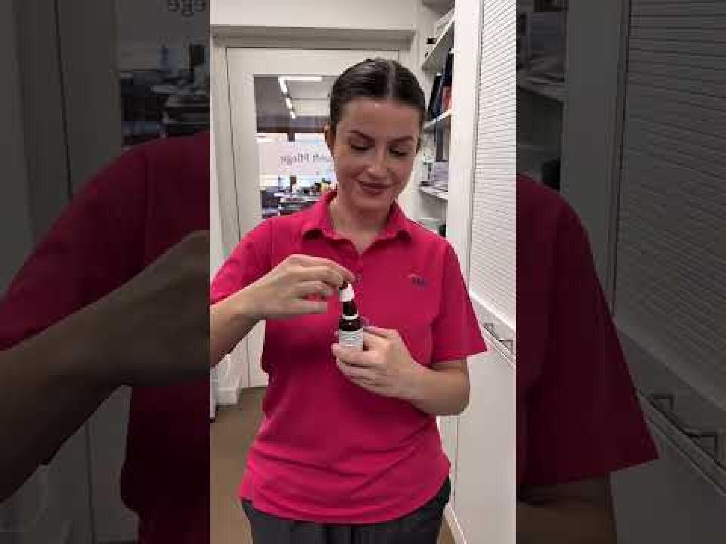 Eine Frau in einem hellrosa Polohemd steht in einem Raum, schaut nach unten und benutzt eine Tropfflasche. Sie scheint sich in einer Apotheke oder einer medizinischen Einrichtung zu befinden, mit Regalen und Schränken im Hintergrund.