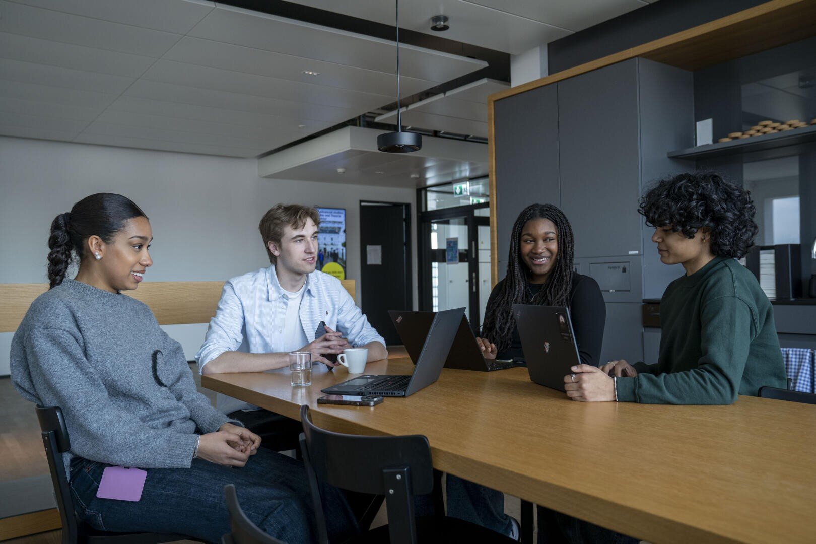 Vier Personen sitzen in einem modernen Büro um einen Tisch herum, unterhalten sich und arbeiten an ihren Laptops. Zwei Frauen und zwei Männer scheinen in eine gemeinsame Diskussion vertieft zu sein. Im Hintergrund des Raums ist ein moderner Küchenbereich zu sehen.