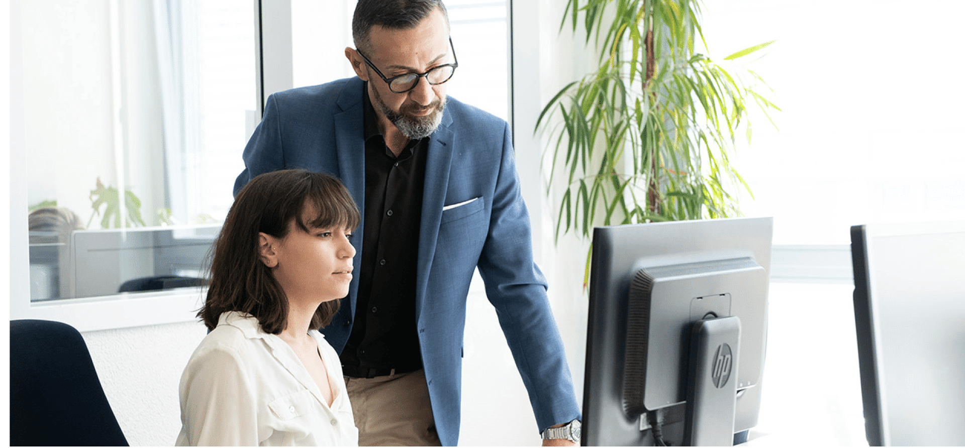 Ein Mann in einem blauen Blazer steht neben einer sitzenden Frau, die beide auf einen Computerbildschirm in einem hellen Büro mit großen Fenstern und einer hohen Pflanze im Hintergrund schauen.