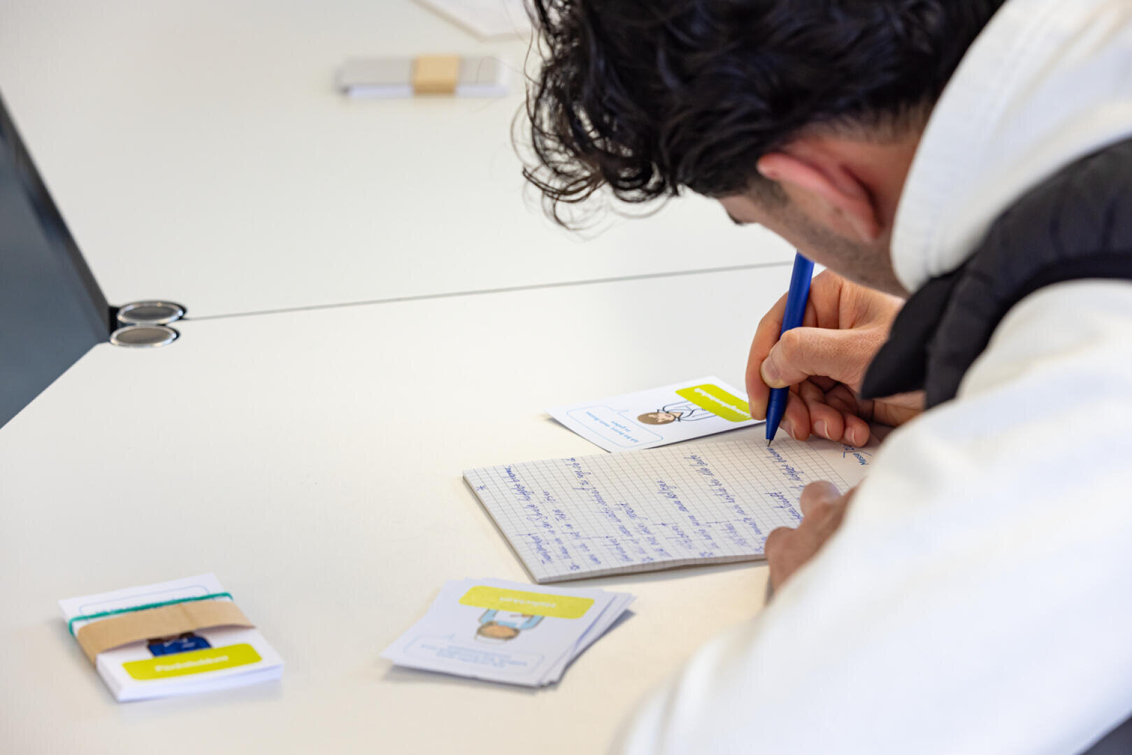 Eine Person mit dunklem Haar schreibt an einem weißen Schreibtisch Notizen in ein Notizbuch. In der Nähe liegen Karten und Papiere, einige mit Illustrationen und Text, ordentlich auf der Oberfläche angeordnet.