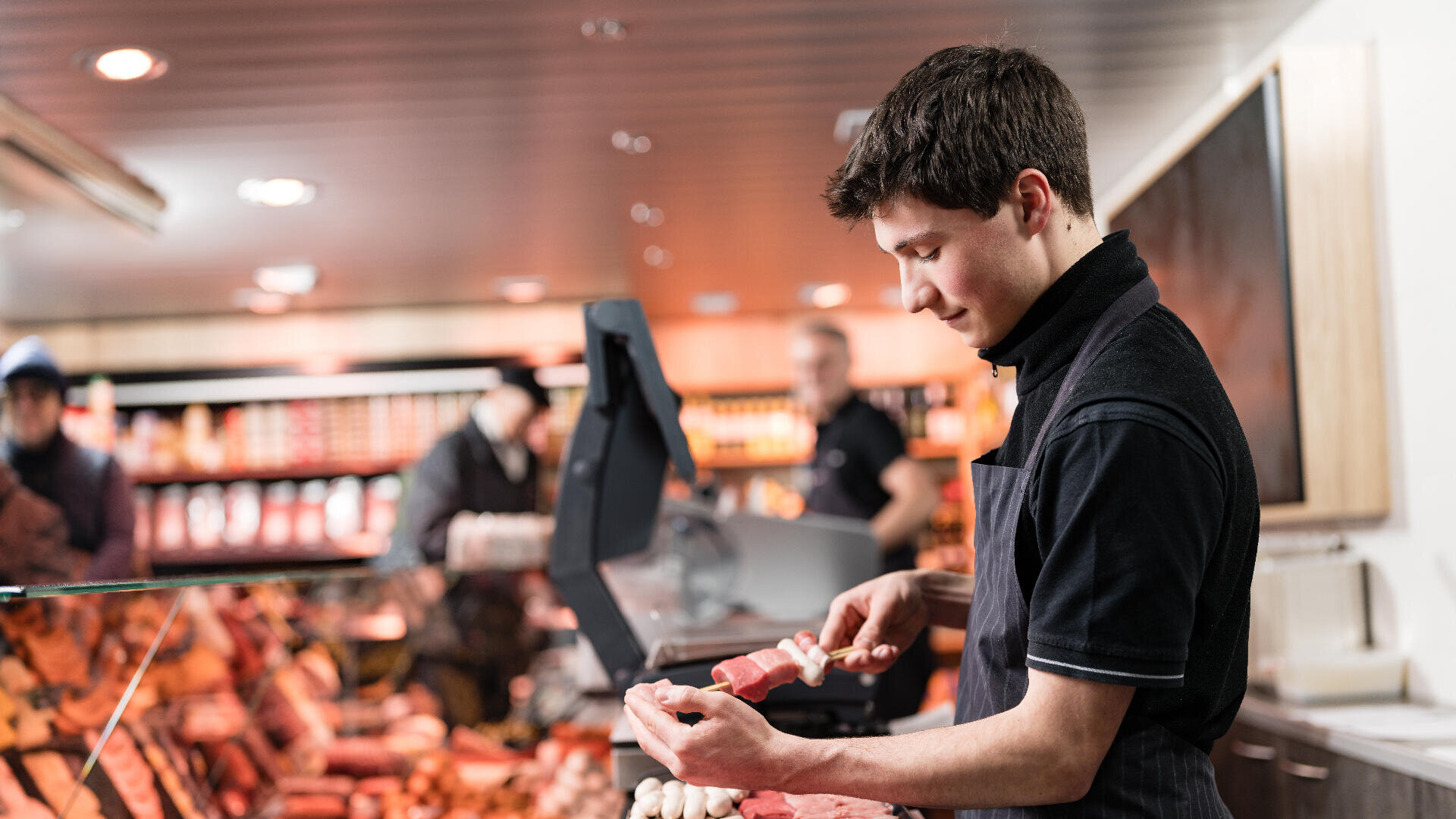 Ein junger Mann in schwarzer Uniform schneidet Fleisch an einer Metzgertheke in einem Geschäft in Scheiben, während im Hintergrund verschiedene Fleischsorten ausgestellt sind und andere Mitarbeiter zu sehen sind.