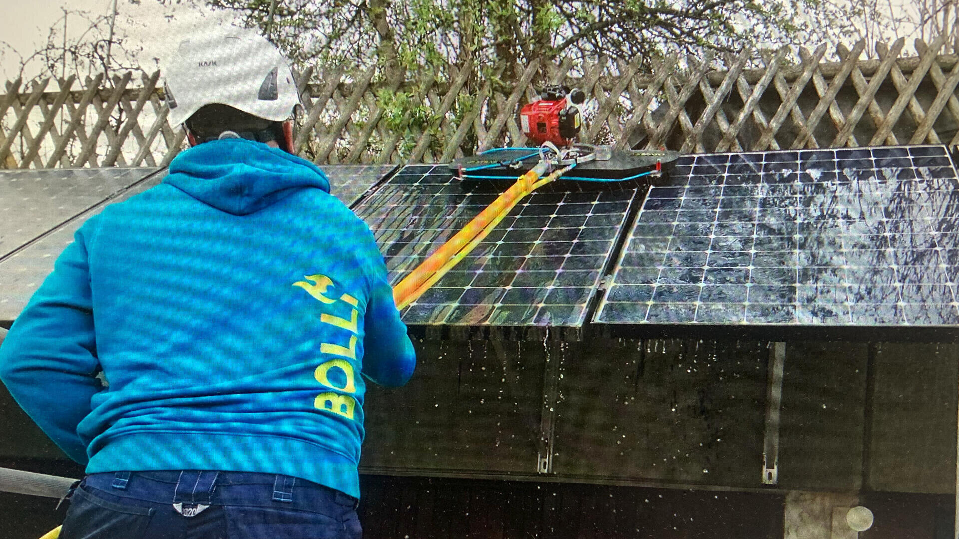 Eine Person in einem blauen Kapuzenpullover und einem weißen Helm benutzt eine lange Stange und eine Maschine, um Wasser von Solarmodulen auf einem Dach zu entfernen. Im Hintergrund sind Bäume und ein Holzzaun zu sehen.