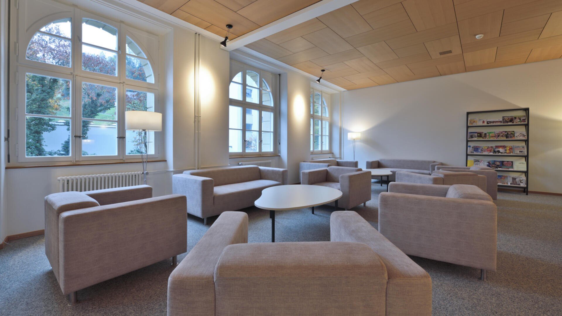 Ein modernes Wohnzimmer mit beigen Sesseln und runden Tischen, die in einem Kreis angeordnet sind, großen Bogenfenstern, die natürliches Licht hereinlassen, und einem Zeitschriftenständer an der Wand. Der Raum hat eine helle Holzdecke und einen neutralen Teppich.