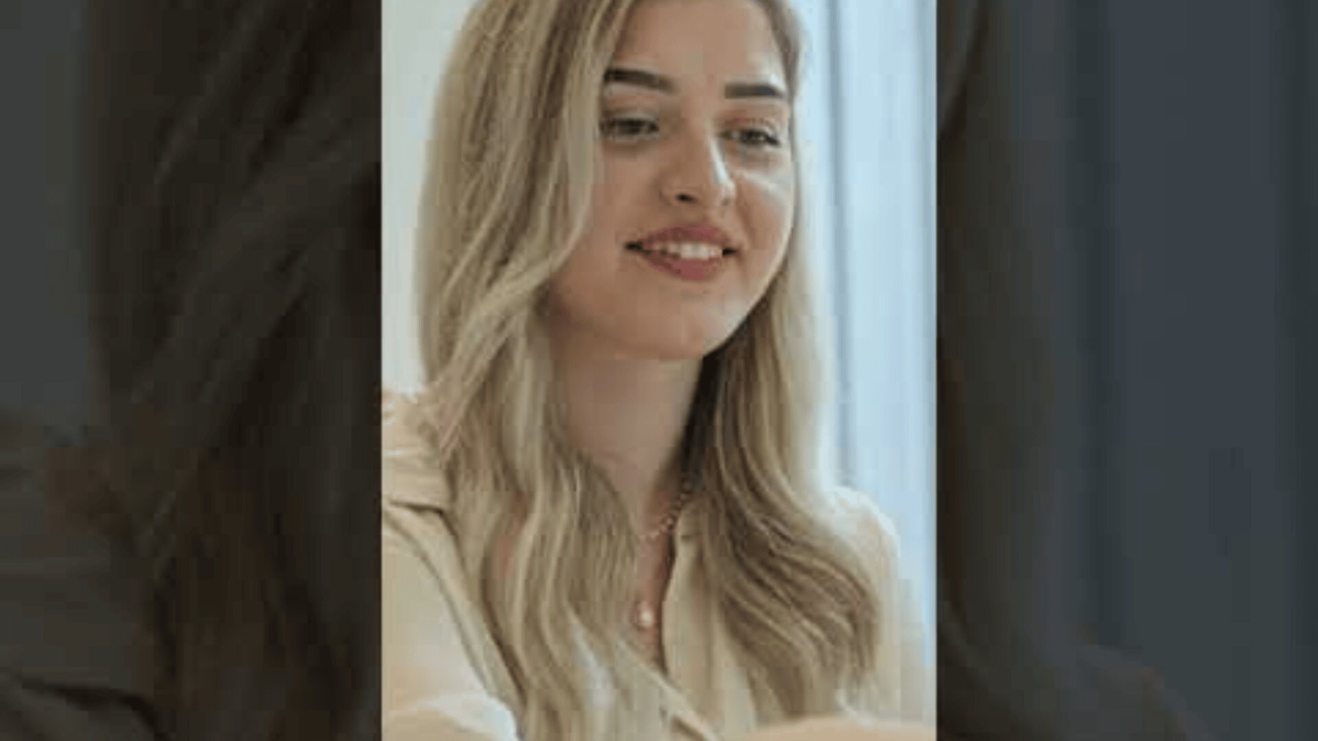 Eine junge Frau mit langem, gewelltem blondem Haar und heller Haut lächelt sanft. Sie trägt ein helles Oberteil und befindet sich in einem Innenraum mit weicher Beleuchtung und einem unscharfen Hintergrund.