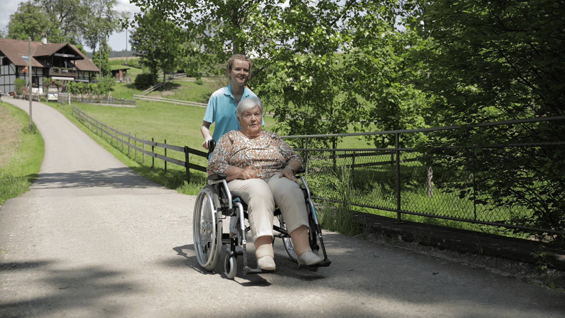 Ein junger Mann schiebt eine ältere Frau im Rollstuhl über einen gepflasterten Weg, der von Grünflächen umgeben ist. Im Hintergrund sind an einem sonnigen Tag ein Haus und Bäume zu sehen.