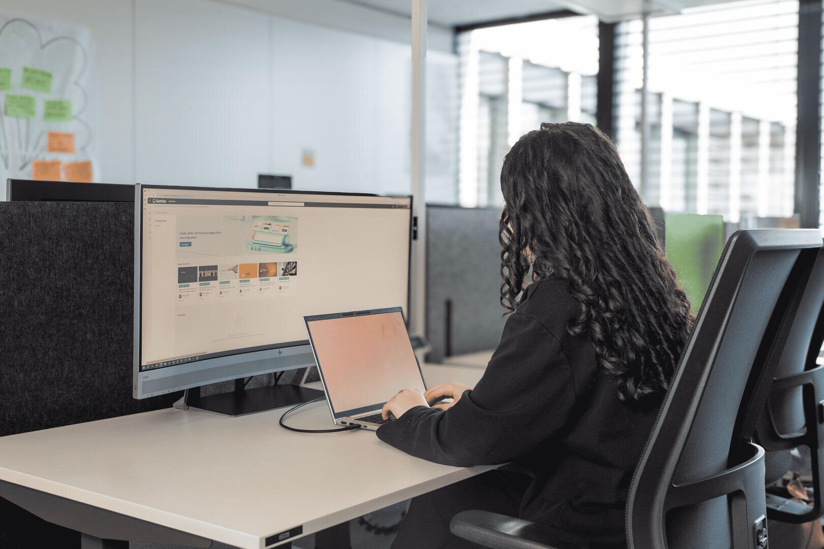 Eine Person mit langen, lockigen Haaren arbeitet an einem Schreibtisch in einem modernen Büro mit einem Laptop, auf dessen großem Bildschirm eine Website zu sehen ist.