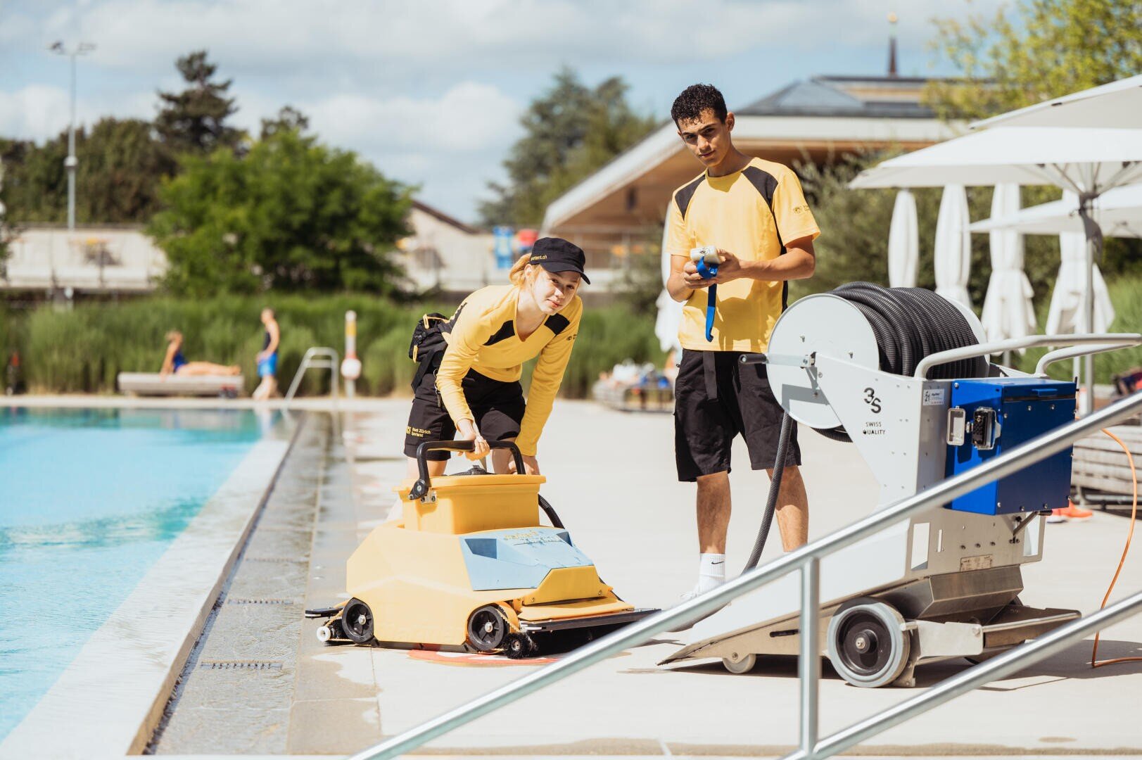 Zwei Arbeiter in gelben Hemden reinigen an einem sonnigen Tag den Rand eines Freibads mit Reinigungsgeräten. Einer bedient einen Bodenschrubber, der andere hält einen blauen Schlauch auf einer Trommel. Im Hintergrund schwimmen Menschen.