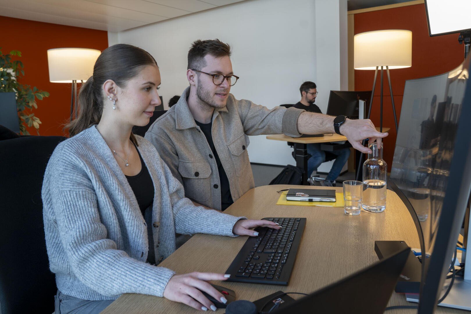 Zwei Personen sitzen gemeinsam an einem Schreibtisch und arbeiten am Computer, wobei eine von ihnen auf den Monitor zeigt. Im Hintergrund arbeitet eine weitere Person an einem Schreibtisch. Das Büro ist modern eingerichtet und verfügt über eine warme Beleuchtung.