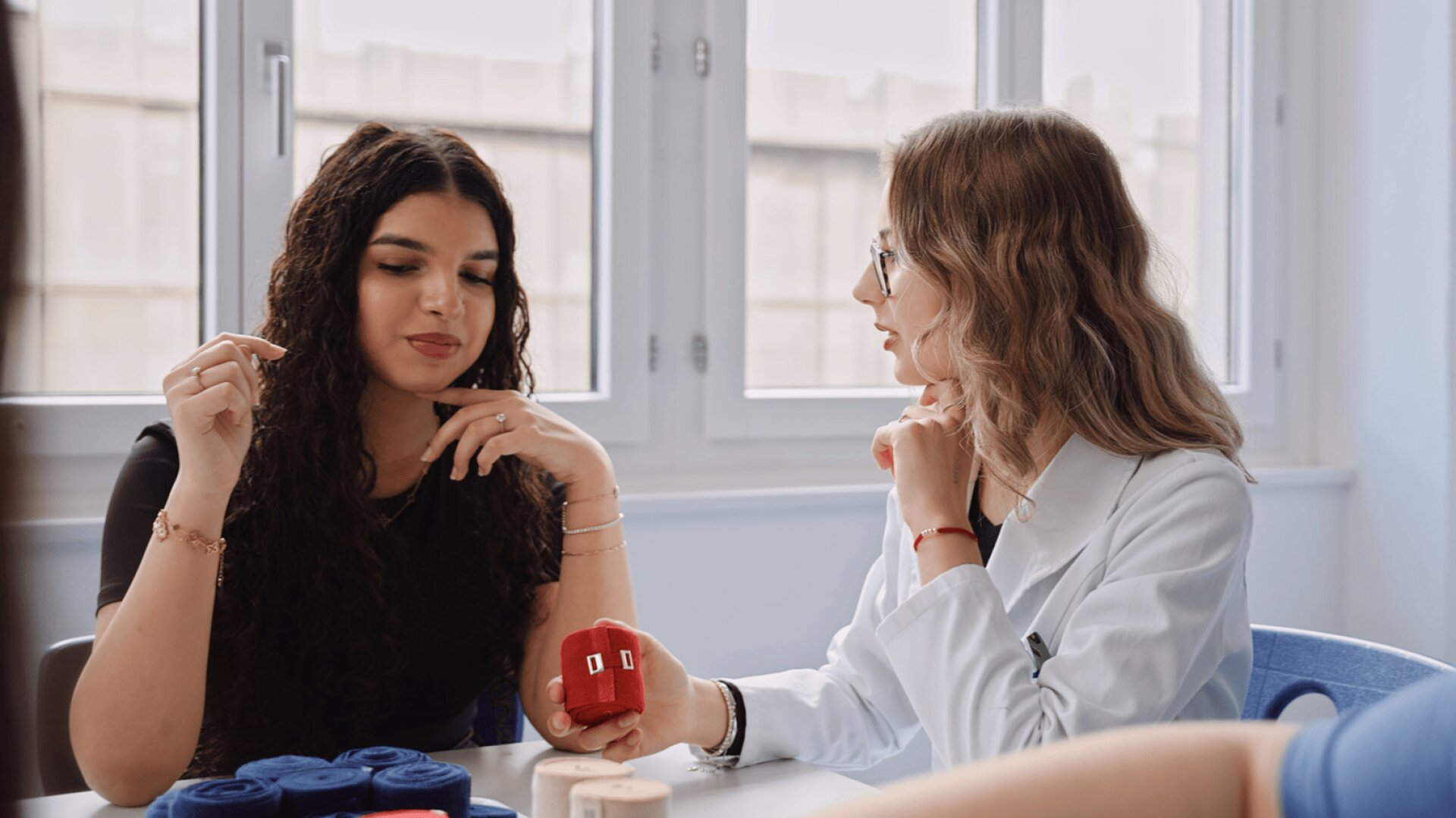 Zwei Frauen sitzen an einem Tisch am Fenster; eine trägt einen weißen Kittel und eine Brille und hält ein rotes Modellherz, während die andere aufmerksam zuhört. Auf dem Tisch liegen Rollen mit Garn oder medizinischen Modellen.