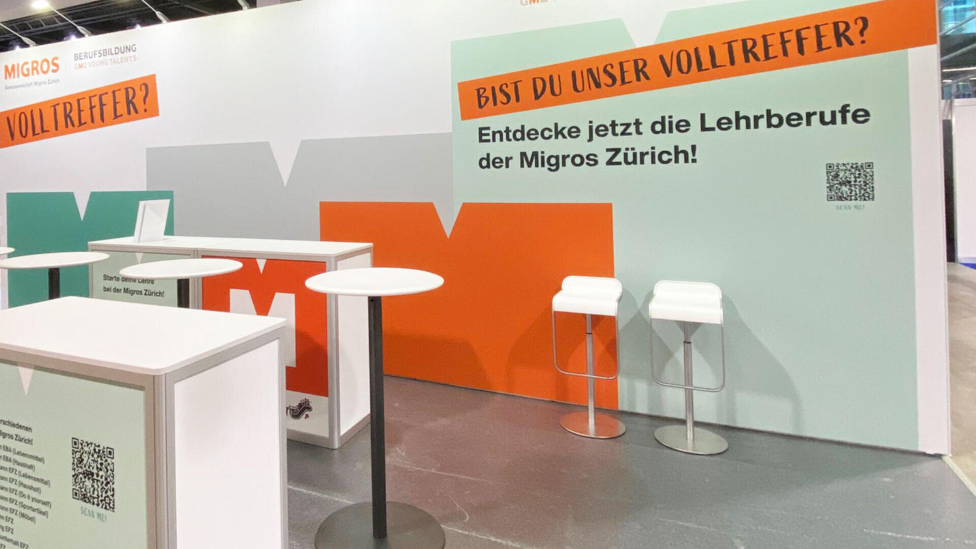 Messestand mit orange-grauen Akzenten, Theke, zwei weissen Hockern und Hinweisschildern auf die Lehrstellen der Migros Zürich. Text und QR-Codes laden die Besucher ein, sich über Karrieremöglichkeiten zu informieren.