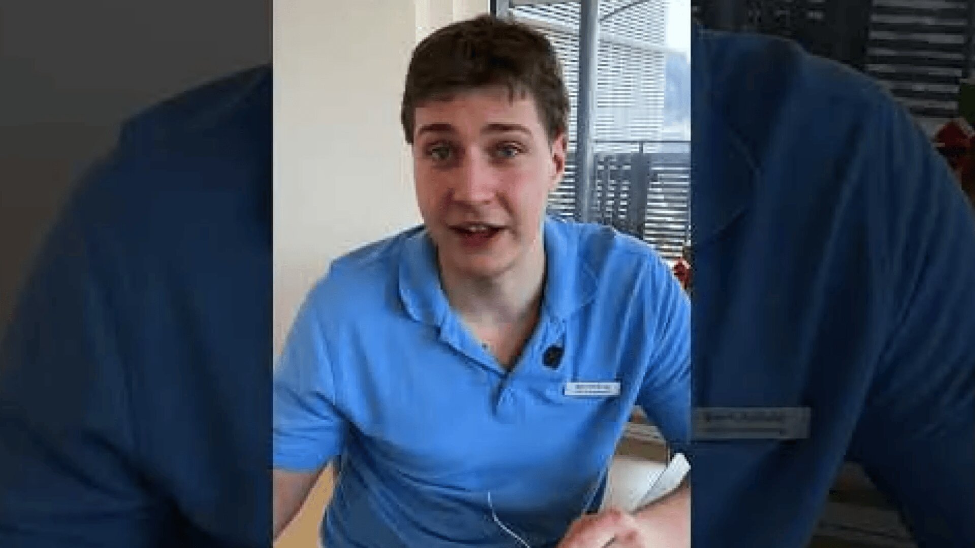Ein junger Mann mit kurzen braunen Haaren, der ein hellblaues Polohemd und ein Namensschild trägt, sitzt in einem Haus und spricht in Richtung der Kamera. Im Hintergrund ist ein Fenster mit Jalousien zu sehen.