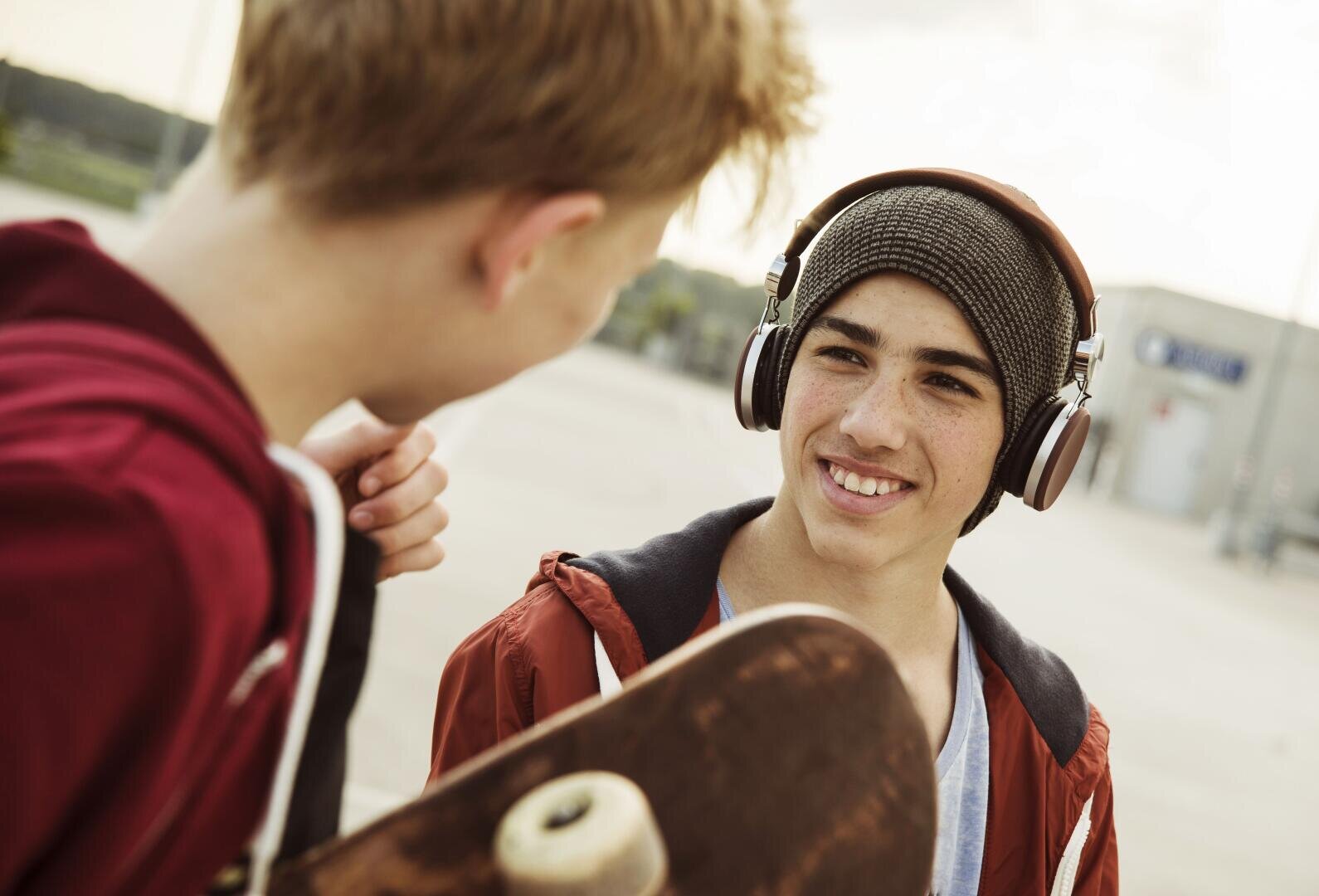 Zwei Teenager in Kapuzenjacken unterhalten sich; einer hält ein Skateboard in der Hand und trägt Kopfhörer.
