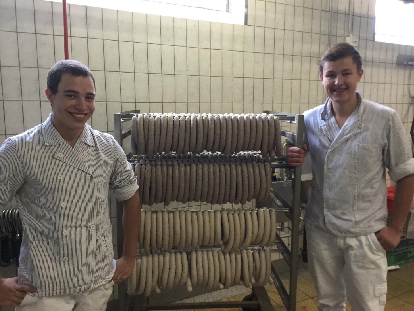 Zwei junge Leute in weißen Uniformen stehen lächelnd neben Regalen mit Reihen hängender Würste in einem gekachelten Raum, wahrscheinlich eine Metzgerei oder Wurstfabrik.