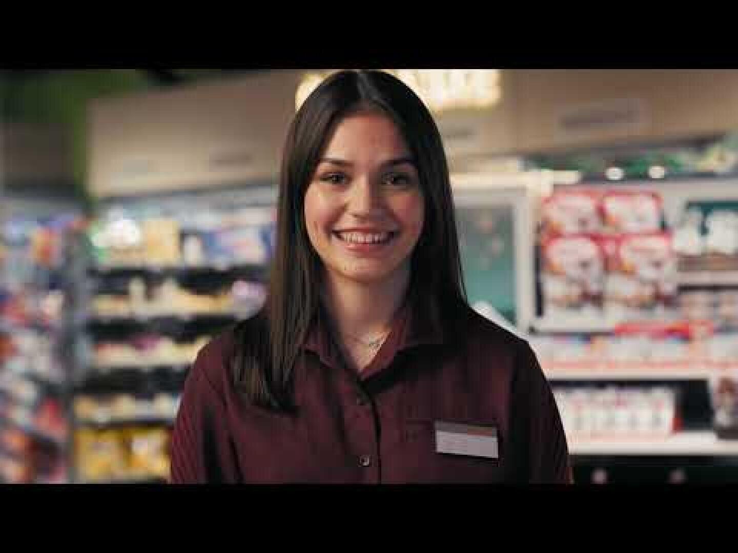 Eine junge Frau mit glattem braunem Haar, die ein kastanienbraunes Uniformhemd und ein Namensschild trägt, lächelt, während sie in einem hell erleuchteten Gang eines Lebensmittelgeschäfts steht. Im Hintergrund sind Regale mit verschiedenen Produkten zu sehen.