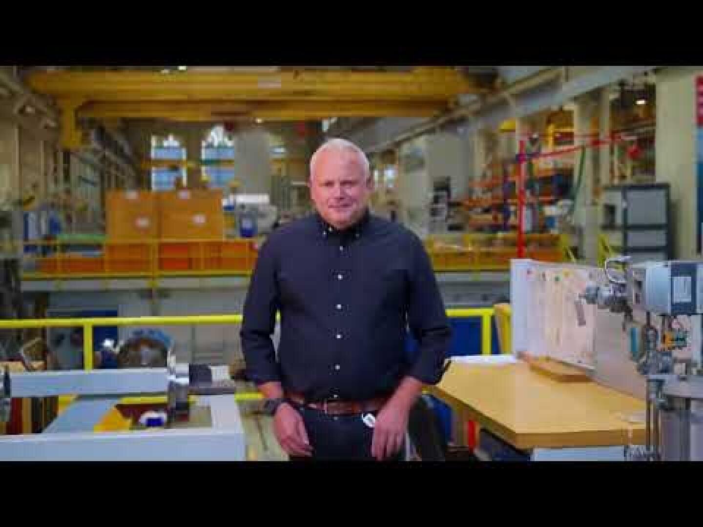 Ein Mann in einem dunklen Hemd steht in einer industriellen Werkstatt oder Fabrik, umgeben von Maschinen, Geräten und Werkbänken, mit Regalen und gelben Sicherheitsgeländern im Hintergrund.