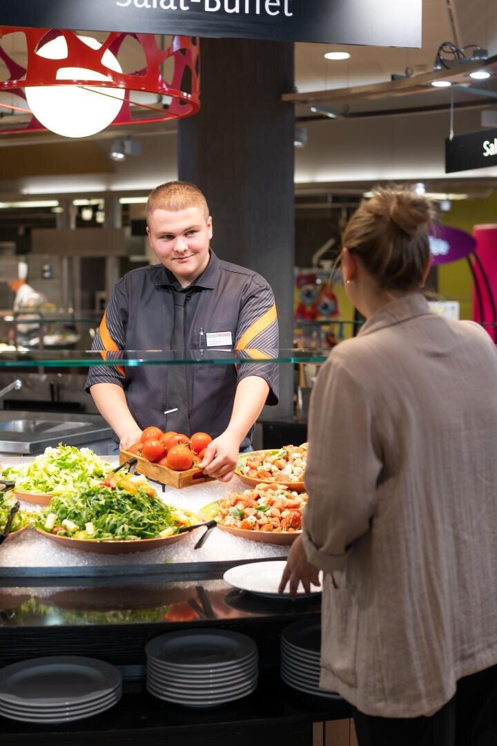 Ein Mann hinter der Theke einer Salatbar reicht einer Frau, die einen Teller in der Hand hält, Tomaten, während unter einem Schild mit der Aufschrift „Salat-Buffet“ verschiedene Salate und Gerichte ausgestellt sind.