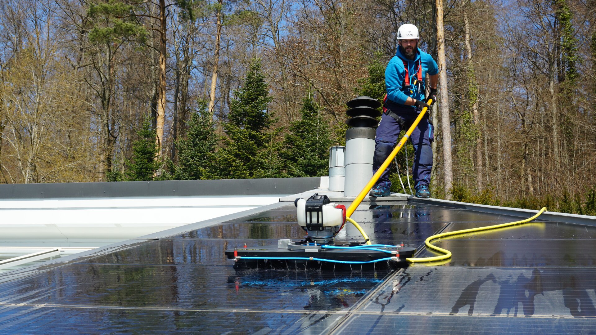 An einem sonnigen Tag wäscht ein Arbeiter in Schutzausrüstung mit einem Reinigungsgerät mit langem Griff Solarmodule auf einem Dach, das von Bäumen und Entlüftungsrohren umgeben ist.