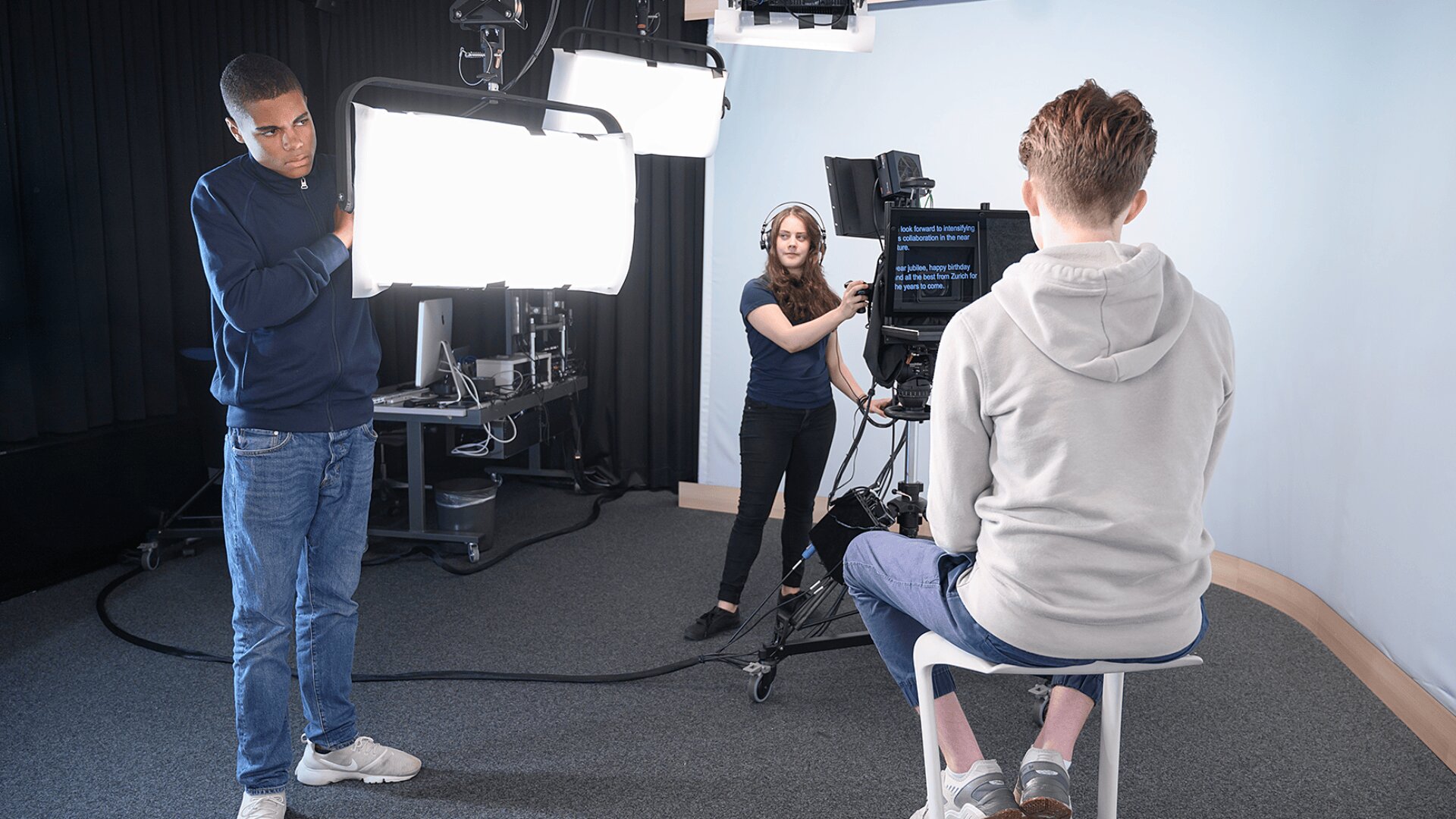 Eine Person sitzt auf einem Hocker und liest von einem Teleprompter ab, während eine Frau die Kamera bedient und ein Mann ein großes Studiolicht in einer professionellen Videoproduktionsumgebung einstellt.