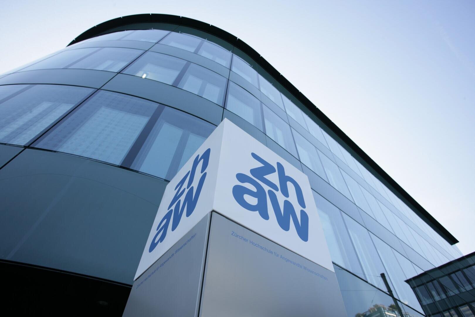 Modernes Glasgebäude mit einem weißen Schild an der Vorderseite, auf dem das blaue „zhaw“-Logo zu sehen ist, das für die Zürcher Hochschule für Angewandte Wissenschaften steht. Die Aufnahme zeigt eine Aufwärtsperspektive, in der die geschwungene Ecke des Gebäudes und die spiegelnden Fenster zu sehen sind.