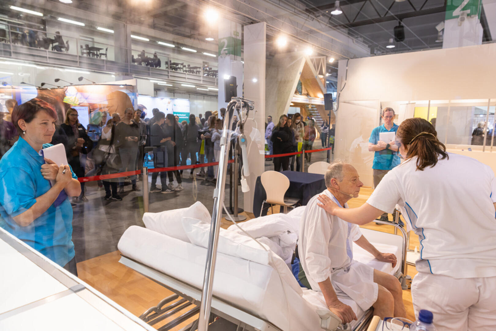 Eine medizinische Simulation: Eine Krankenschwester untersucht einen älteren Mann, der in einem Krankenhausbett liegt, während eine andere Mitarbeiterin ihn beobachtet und die Leute von außerhalb einer Glaswand in einer belebten Ausstellungshalle zuschauen.