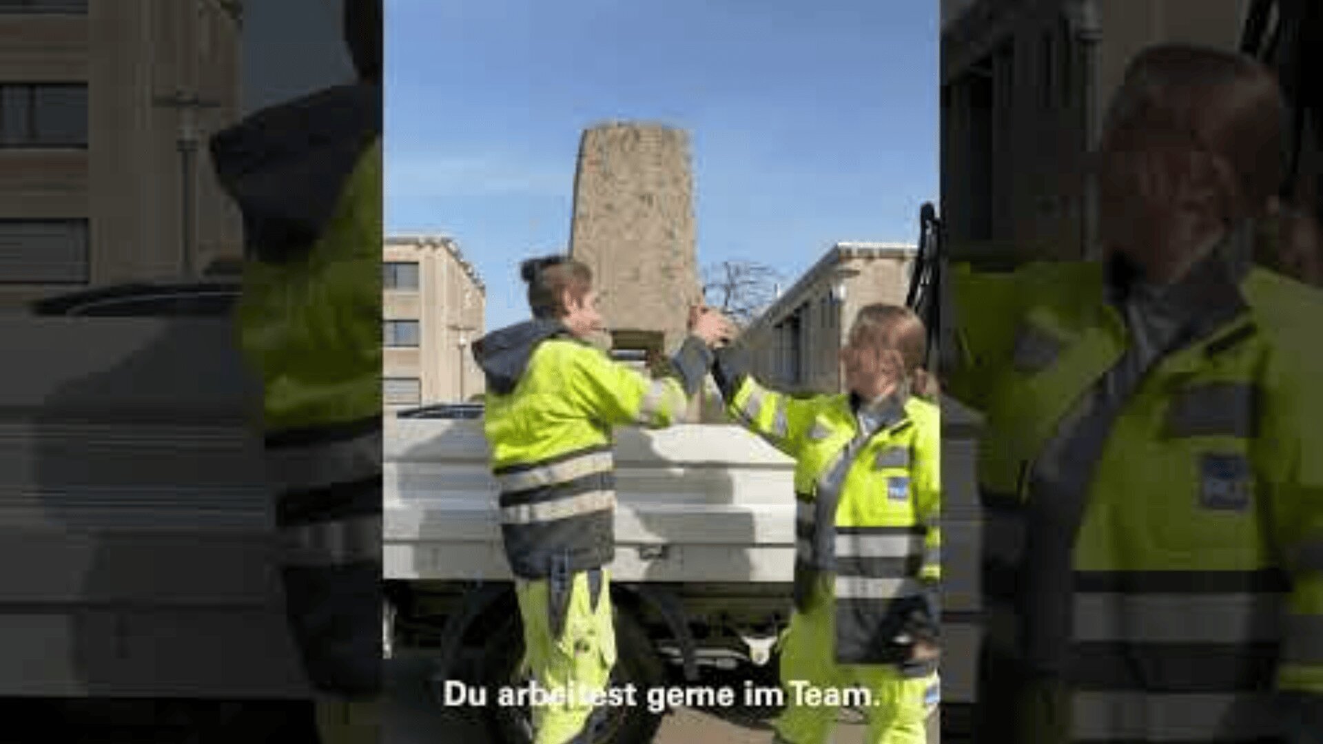 Zwei Arbeiter in gelben Sicherheitsjacken stehen draußen in der Nähe eines Lastwagens und geben sich gegenseitig einen Faustschlag. Im Hintergrund sind Gebäude zu sehen, und der deutsche Text am unteren Rand lautet: "Du arbeitest gerne im Team".