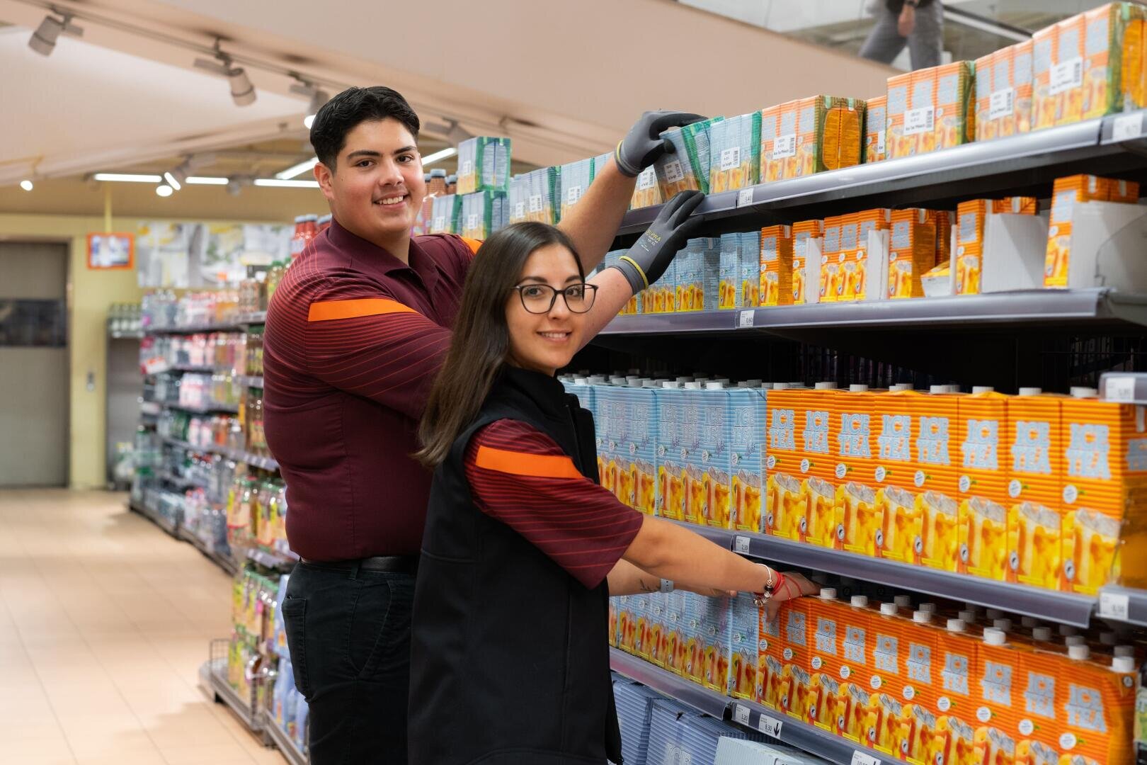 Zwei Supermarktmitarbeiter, ein Mann und eine Frau, füllen die Regale mit Saftkartons auf. Beide tragen Uniformen und Handschuhe und lächeln in die Kamera in einem gut beleuchteten Gang eines Lebensmittelgeschäfts.