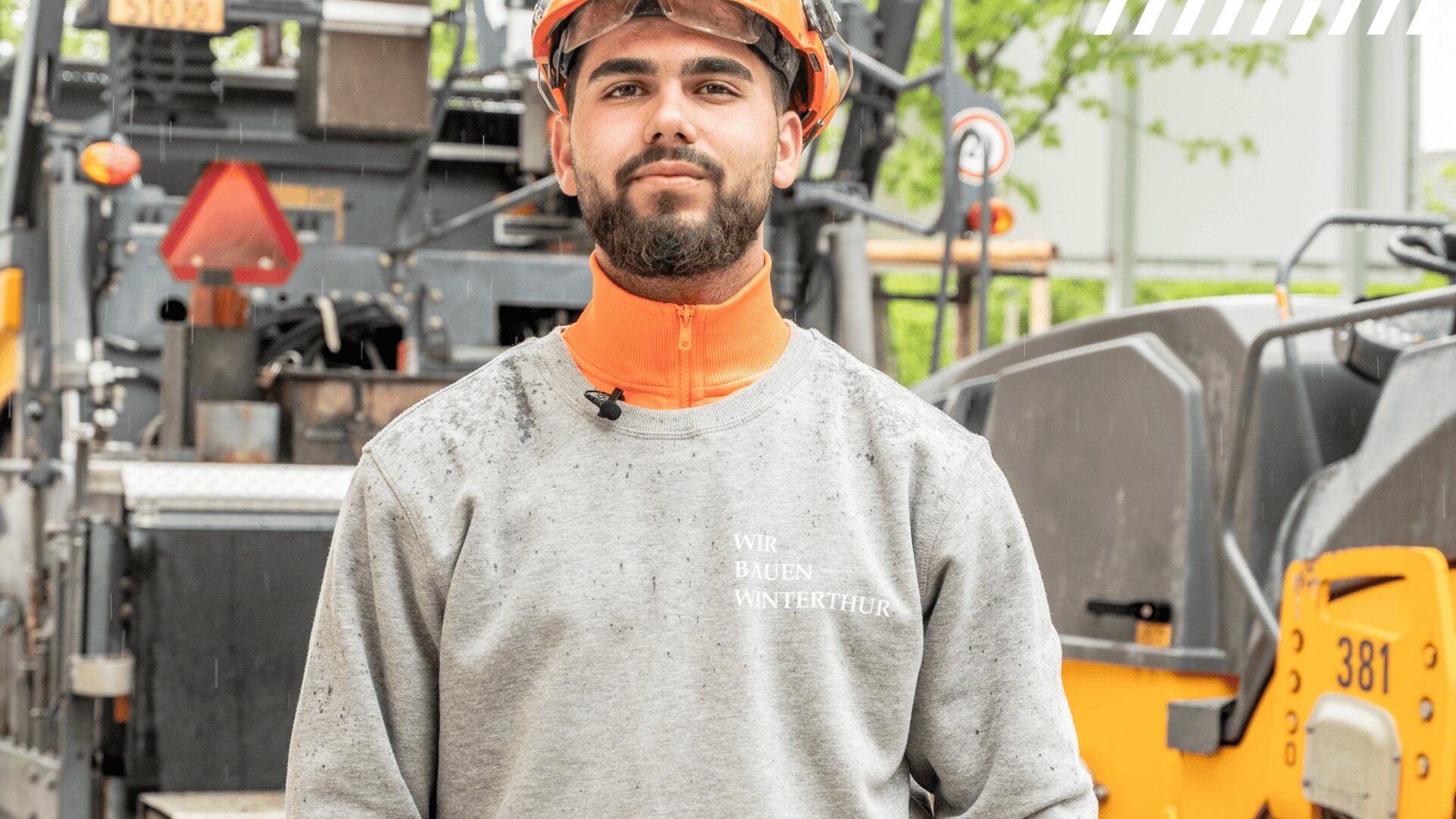 Ein Bauarbeiter, der einen orangefarbenen Helm und einen grauen Pullover trägt, steht auf einer Baustelle vor schweren Maschinen und schaut direkt in die Kamera.