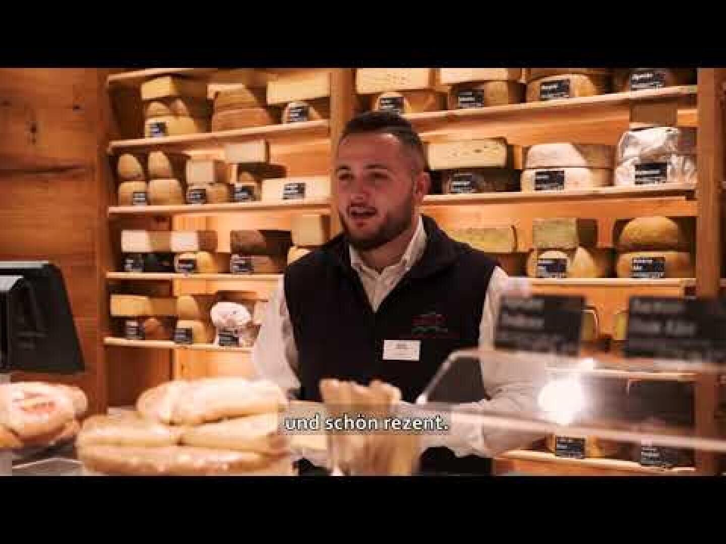 Ein Mann steht hinter einer Theke mit Brot und Gebäck und vor Regalen mit Käse. Er trägt eine dunkle Weste und ein Namensschild. Der Untertitel lautet und schon.ireznt.