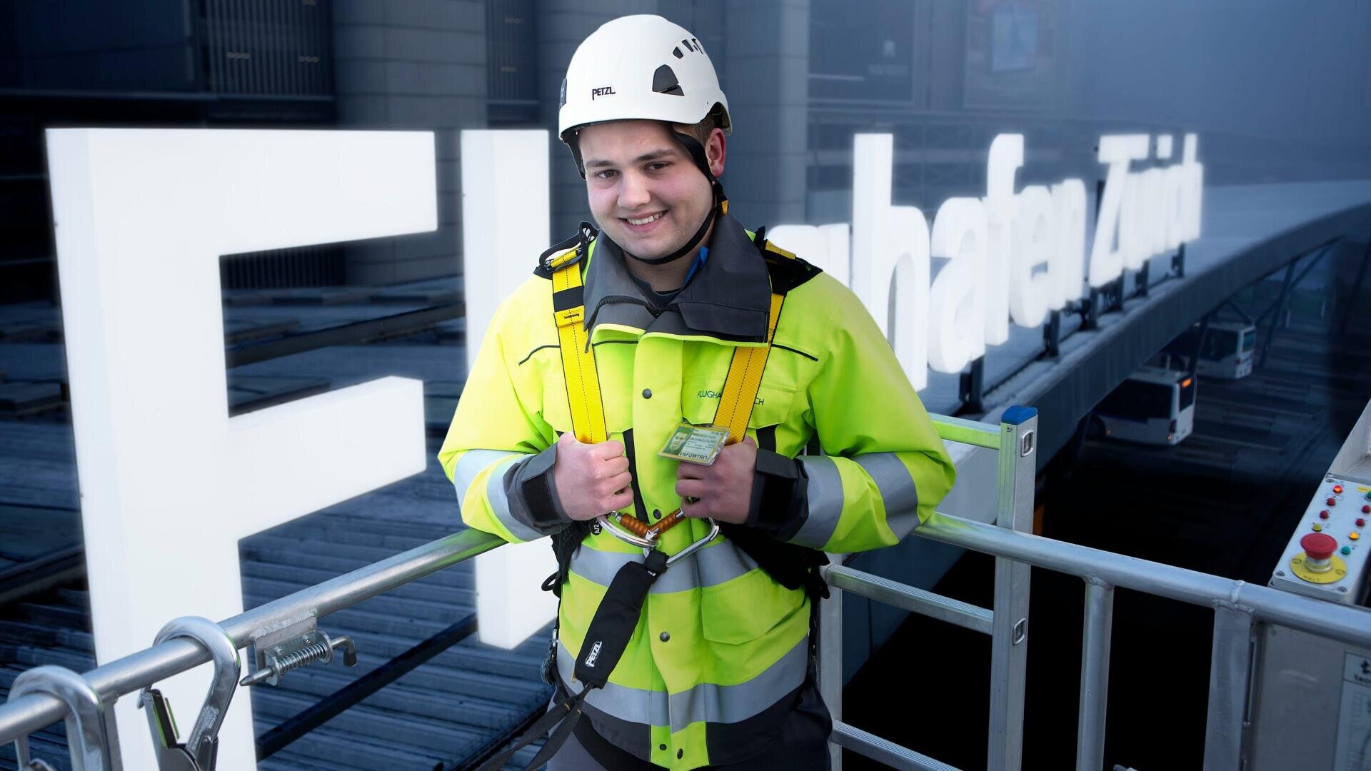 Ein lächelnder Arbeiter mit Schutzhelm und Warnweste steht auf einer Plattform vor großen Leuchtbuchstaben. Der Hintergrund ist leicht neblig und industriell.
