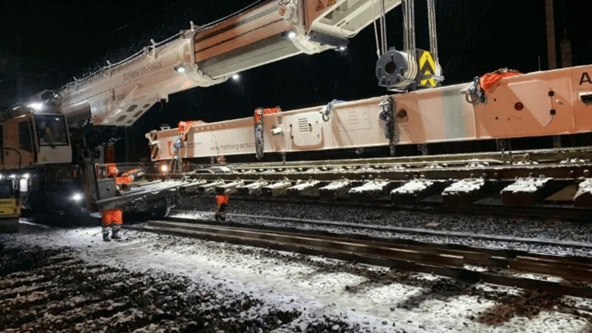 Arbeiter in orangefarbener Schutzkleidung führen einen großen Kran, der nachts, wenn Schnee den Boden und die bestehenden Gleise bedeckt, einen neuen Gleisabschnitt verlegt.