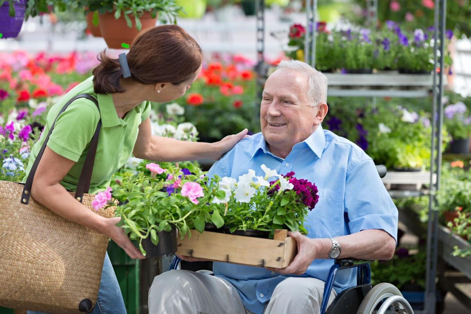 Eine Frau lächelt und unterhält sich mit einem älteren Mann im Rollstuhl, der eine mit bunten Blumen gefüllte Holzkiste in der Hand hält; sie sind umgeben von blühenden Pflanzen in einem Gartencenter.