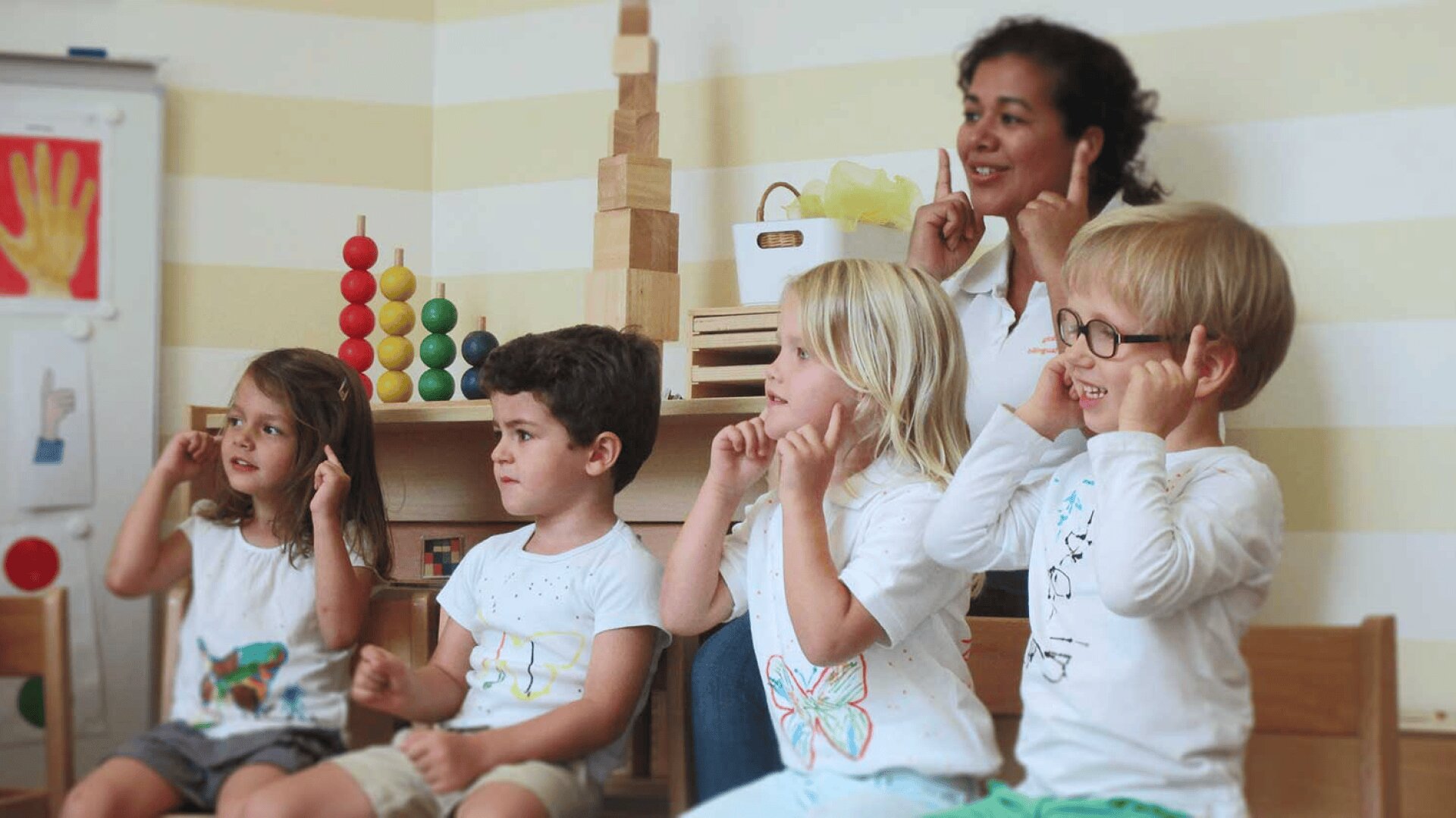 Vier kleine Kinder sitzen in einem Klassenzimmer mit einer Lehrerin, die sich alle die Ohren anfassen und lächeln. Im Hintergrund sind Holzspielzeug und Lernplakate zu sehen. Die Atmosphäre ist spielerisch und einnehmend.