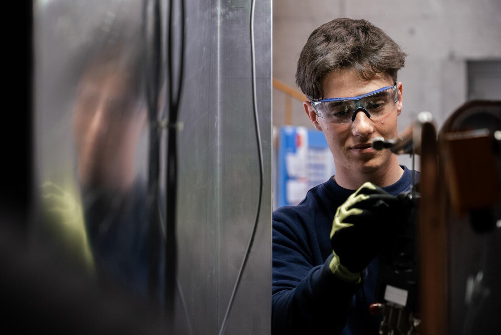 Eine Person, die eine Schutzbrille und Handschuhe trägt, bedient Maschinen in einer Industrie- oder Werkstatteinrichtung, wobei sie sich auf ihre Aufgabe konzentriert und eine reflektierende Oberfläche neben sich hat.