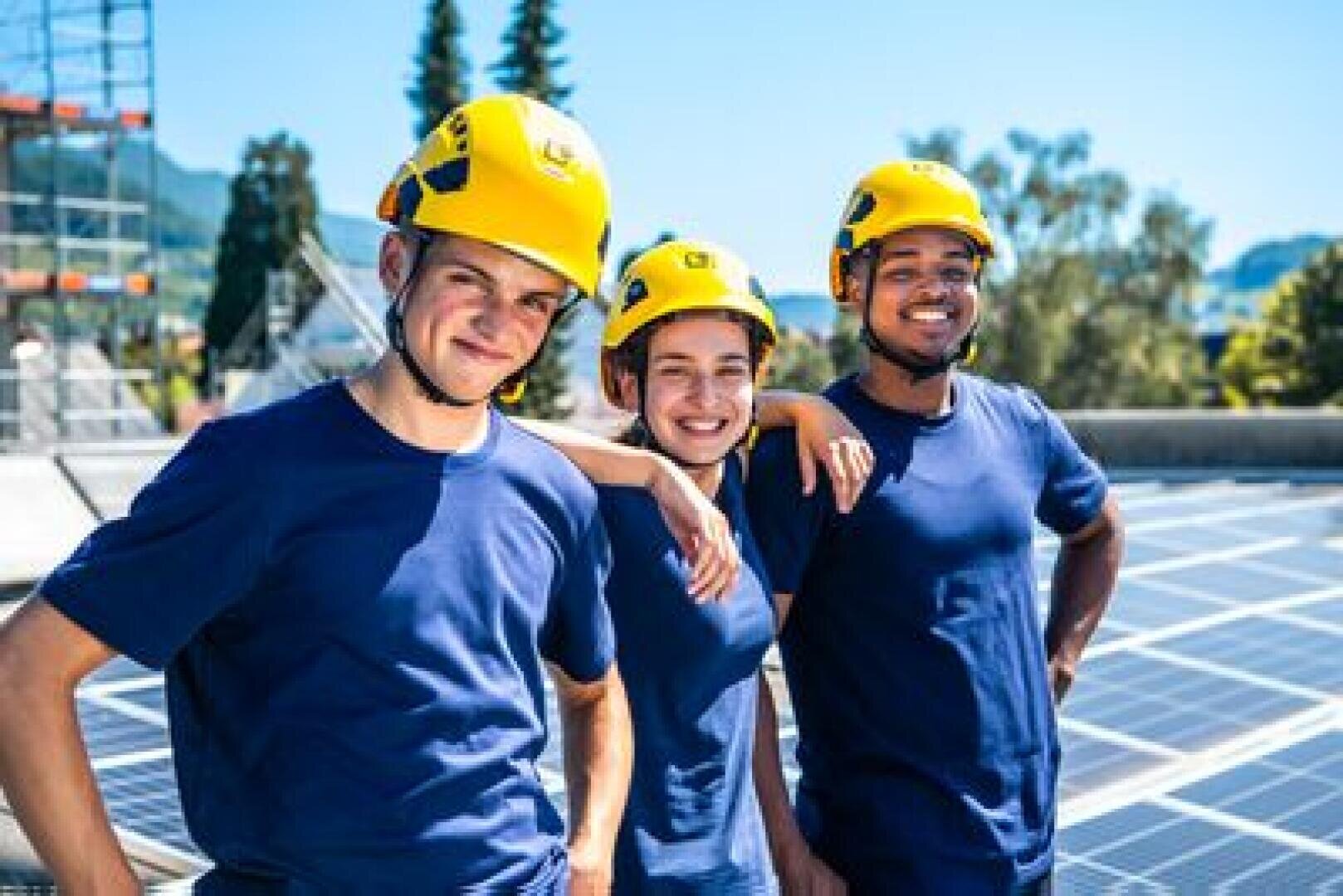 Drei Personen mit gelben Helmen und blauen Hemden stehen an einem sonnigen Tag eng beieinander im Freien und lächeln, während im Hintergrund Sonnenkollektoren und Bäume zu sehen sind.