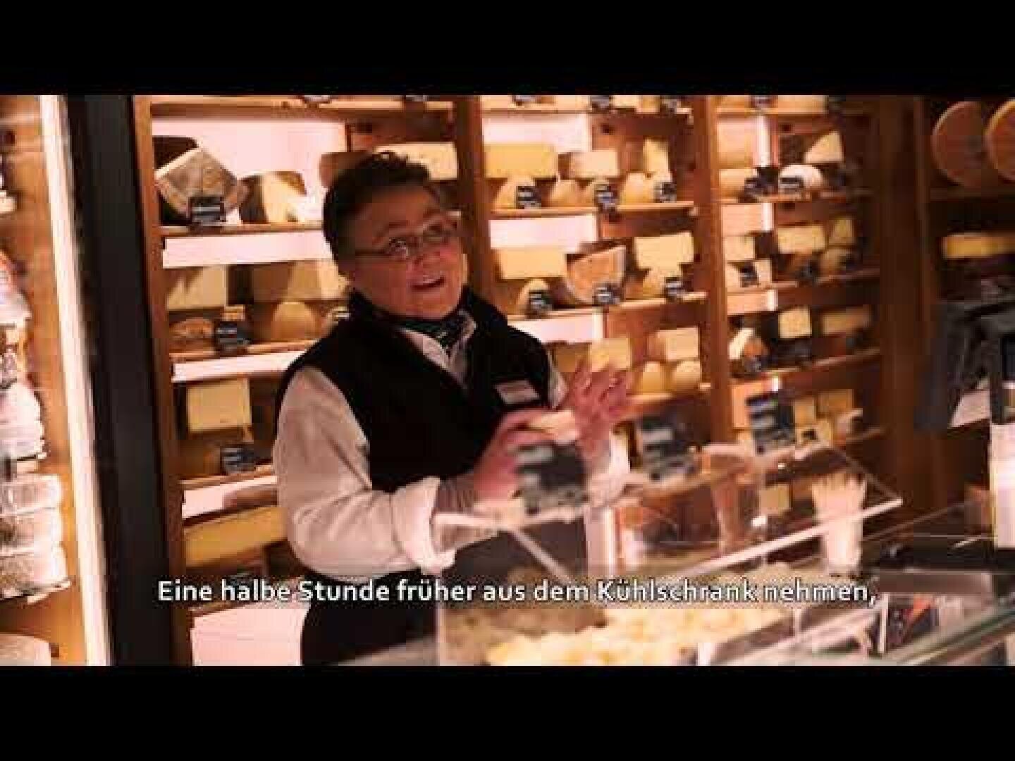 Eine lächelnde Person steht hinter einer gläsernen Käsetheke in einem Geschäft, umgeben von Regalen mit Käse. Die deutschen Untertitel am unteren Rand lauten: "Eine halbe Stunde früher aus dem Kühlschrank nehmen.