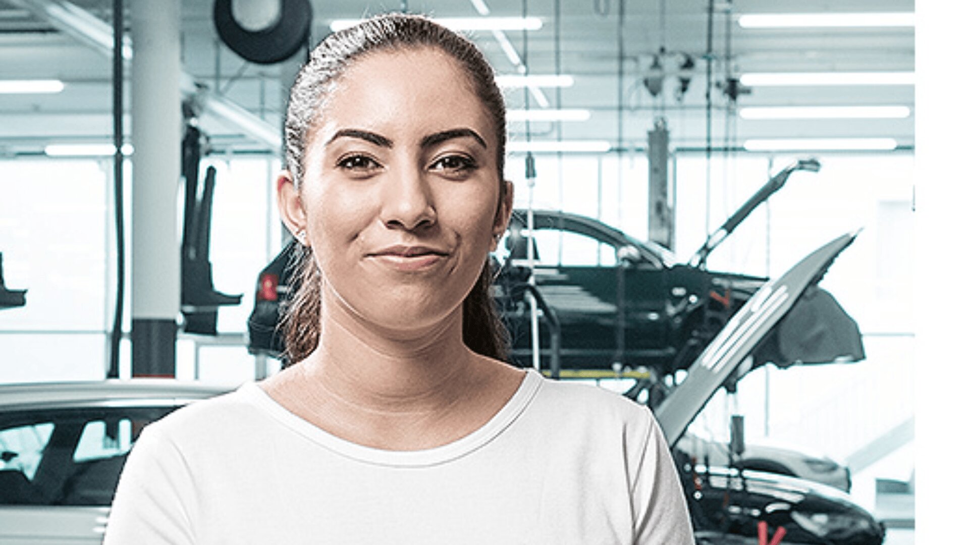 Eine Frau mit zurückgebundenem braunem Haar und weißem Hemd steht in einer modernen Autowerkstatt, in der im Hintergrund Autos und Geräte zu sehen sind. Sie lächelt leicht und schaut in die Kamera.