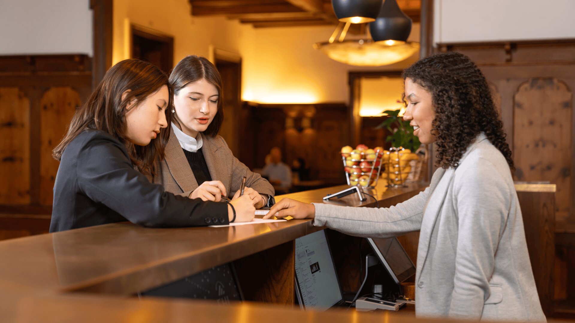 Zwei Frauen stehen an einer Hotelrezeption und checken ein, während eine Rezeptionistin auf ein Dokument zeigt, das sie unterschreiben. Die Umgebung ist warm beleuchtet, mit Holzdekor und einer Schale mit Äpfeln auf dem Tresen.