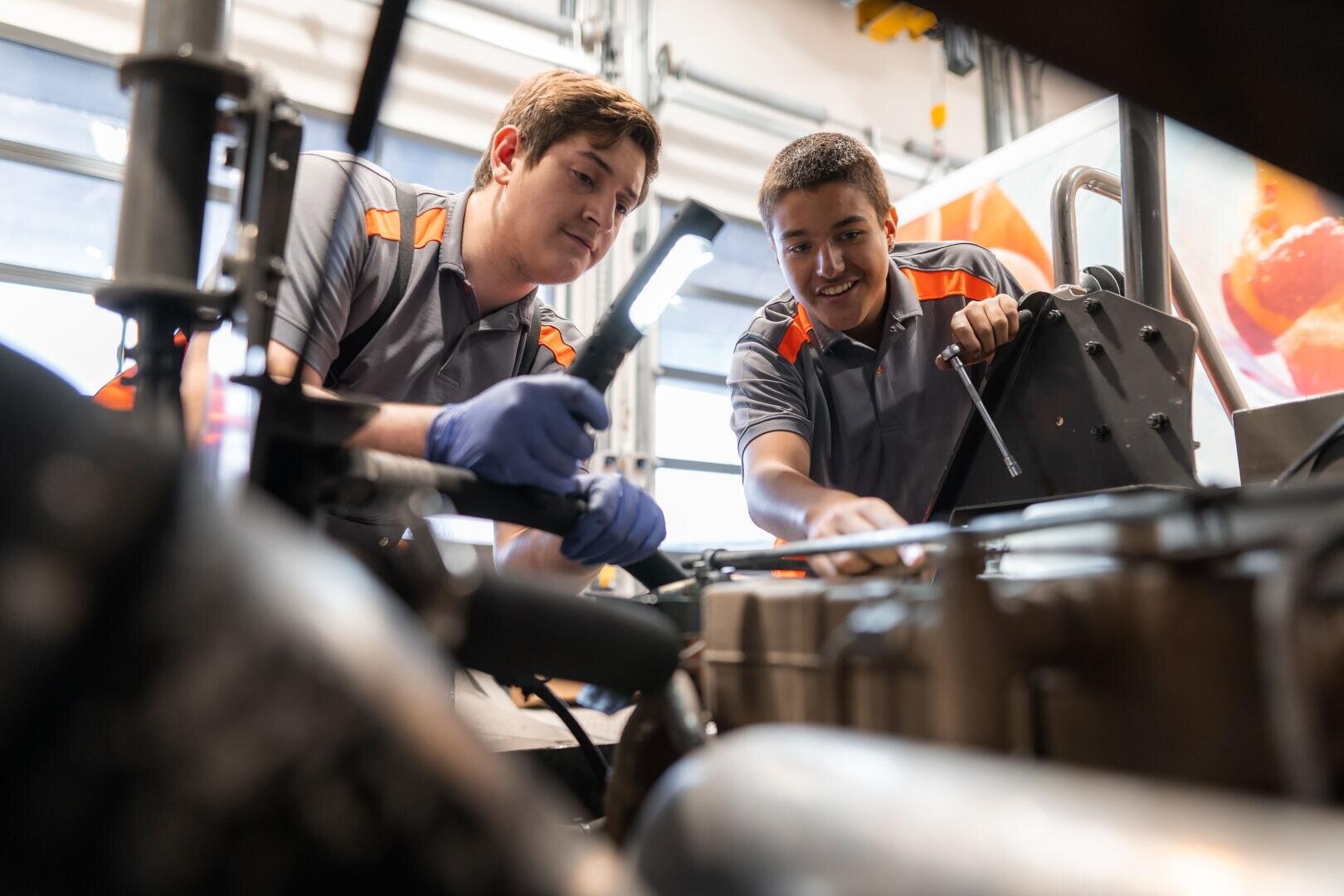 Zwei Mechaniker in grauen Uniformen mit orangefarbenen Akzenten arbeiten gemeinsam an Maschinen, bedienen Werkzeuge und prüfen Teile in einer gut beleuchteten Werkstatt.